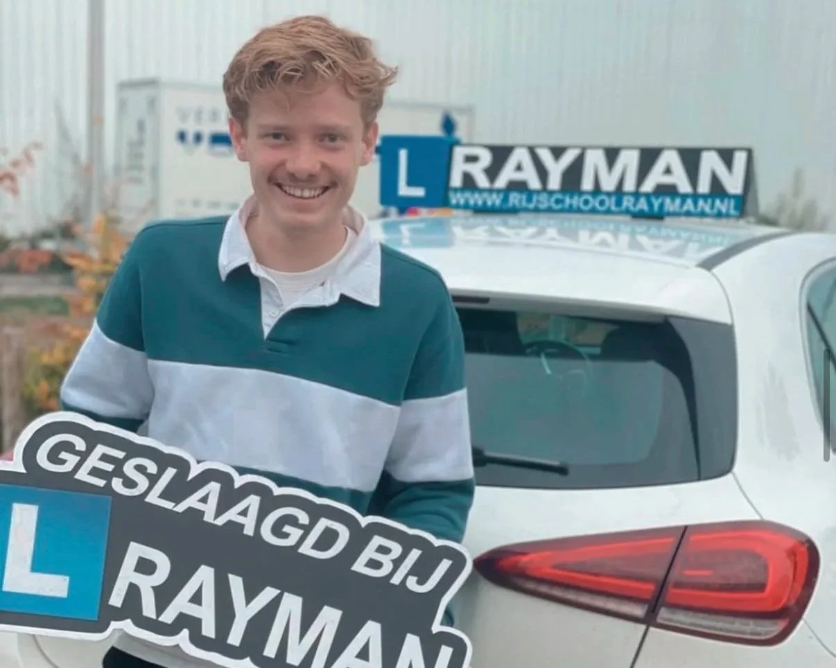 Een lachende jonge man staat voor een auto met een bord dat zegt 'GESLAAGD BIJ L RAYMAN' en een schoolauto met een 'L'-bord en de tekst 'WWW.RJSCHOOLRAYMAN.NL'.