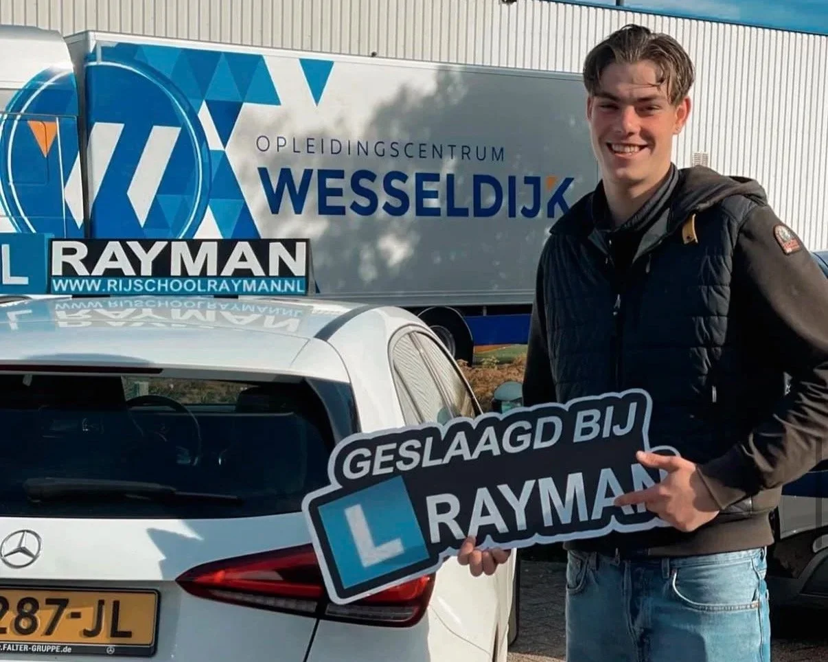 Een jonge man die geslaagd is voor zijn rijbewijs, hij houdt een bord vast met de tekst 'GELUKT GESLAAGD BIJ RAYMAN'. Hij staat naast een witte auto met een rijbewijsnummer en een L-rijbewijs, op de achtergrond een opleidingscentrum met de naam 'WESS
