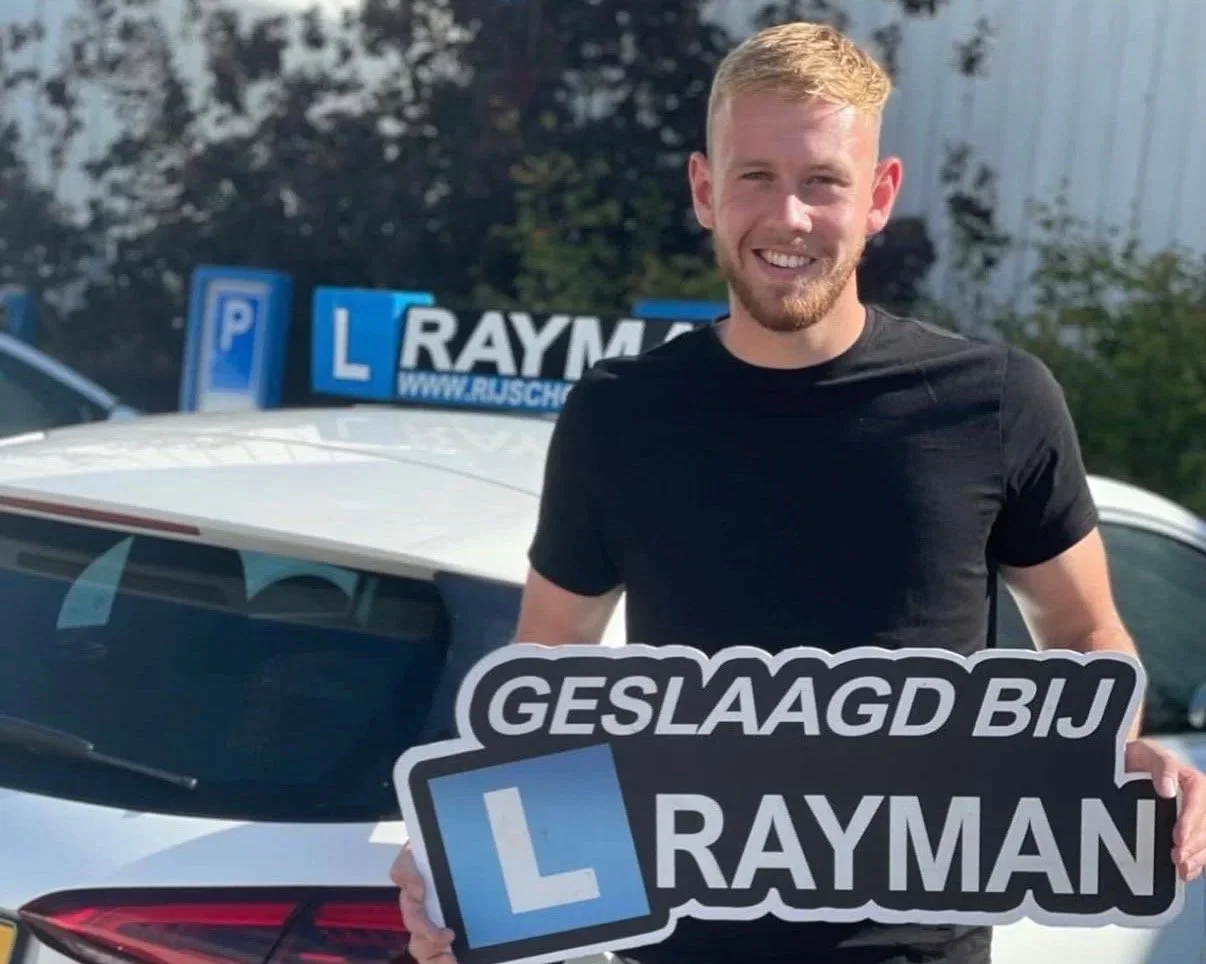 Blije man met blond haar en baard houdt een bord vast dat zegt 'Geslaagd bij Rayman' met een blauwe L. Achter hem staat een auto met een rijopleidingssignaal.