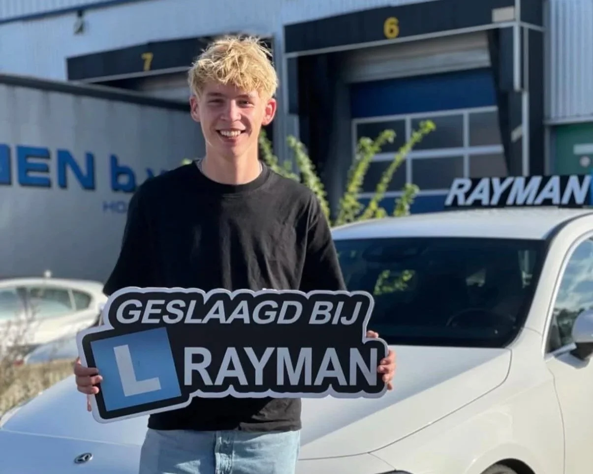 Een jongeman houdt een bord vast met de tekst 'Geslaagd bij Rayman' en een blauw 'L' symbool, terwijl hij voor een witte auto staat. Achtergrond is een autodealer of parking met bedrijfsgebouwen.