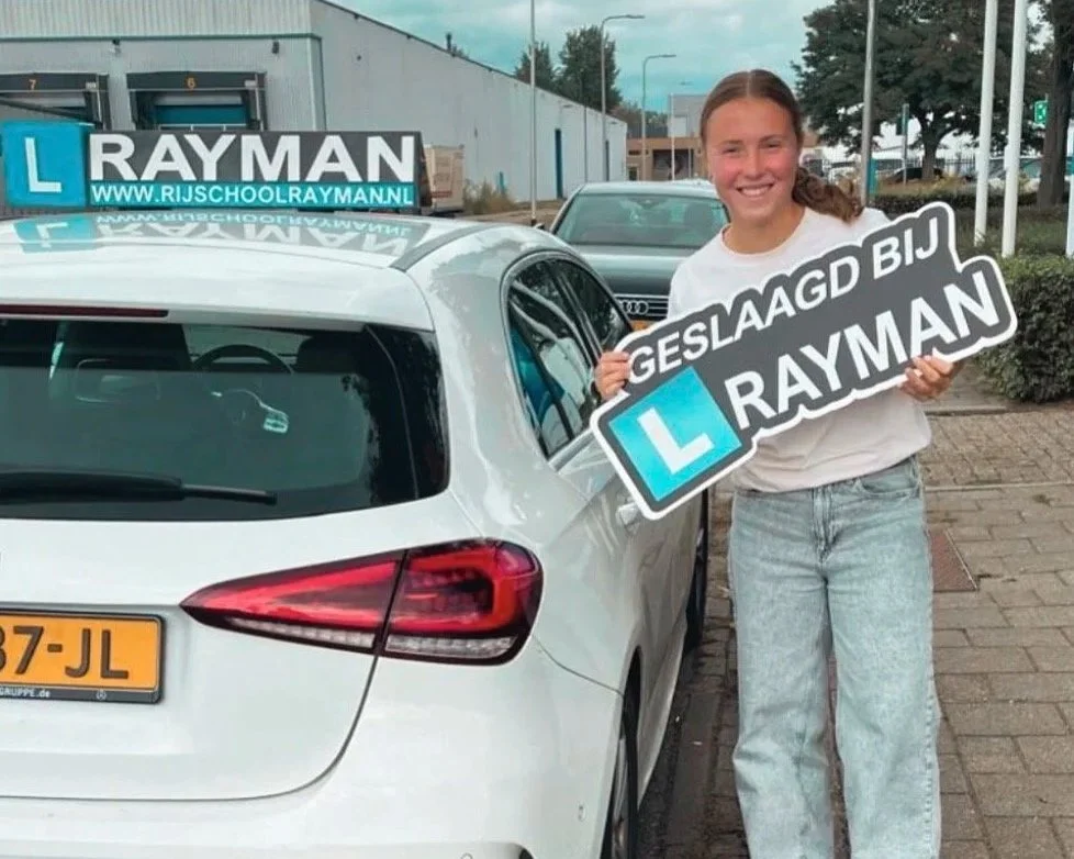 Een meisje staat naast een witte auto en houdt borden vast die zeggen 'Geslaagd bij Rayman' en de website 'www.rijschoolraymann.nl'.