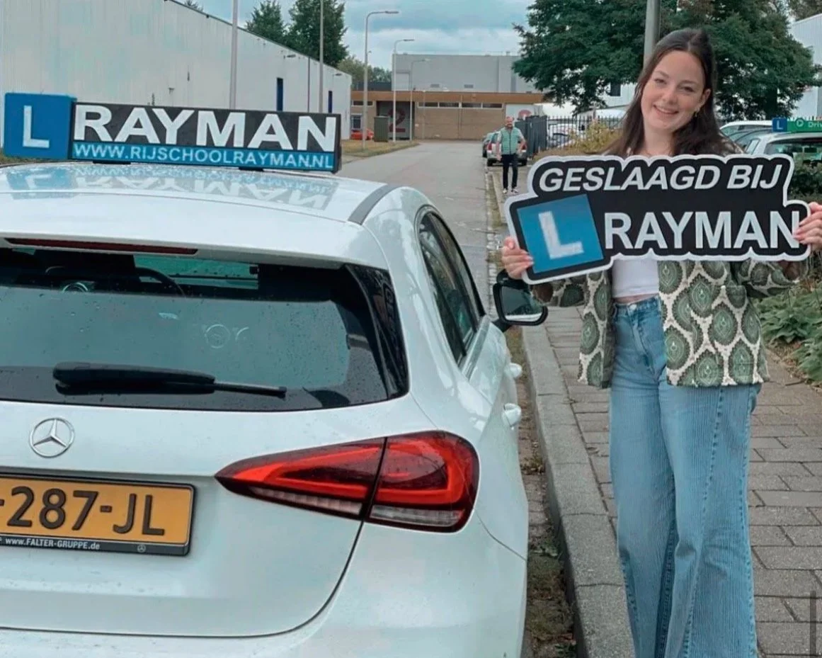 Vrouw met een bord dat zegt 'Geslaagd bij Rayman', vlak achter een witte auto met een inschrijvingsbord. Ze lacht en staat op een stoep.