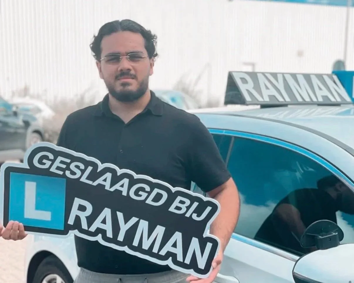 Man houdt een bord met de tekst 'Geslaagd bij Rayman' en een ‘L’ voor rijlessen, voor een auto met een Rayman-taxi op het dak.