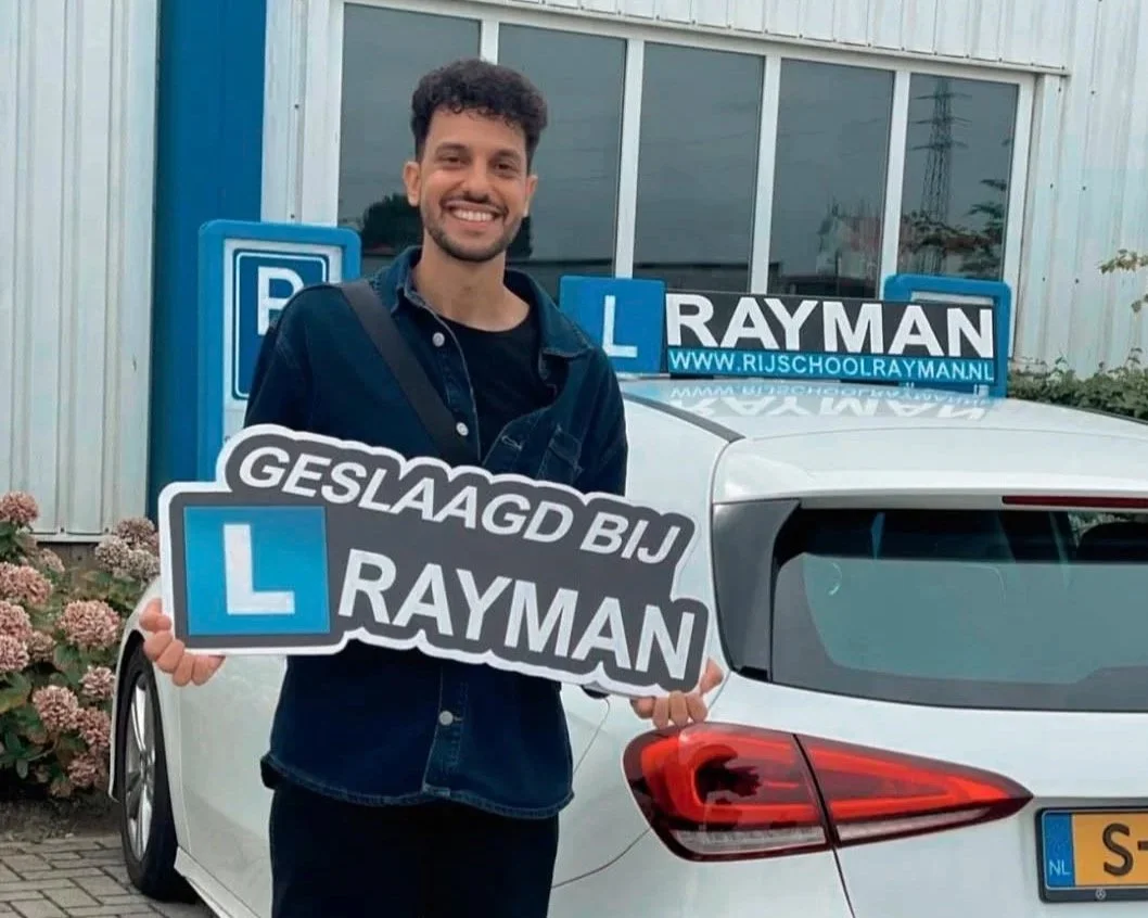 Man staat voor een wit auto met een bord en foto's en tekst op de auto. Hij houdt een bord vast dat zegt 'Geslaagd bij Rayman' met een L-velsign en er staan meer tekens en woorden op de auto en op een bord op de achtergrond.