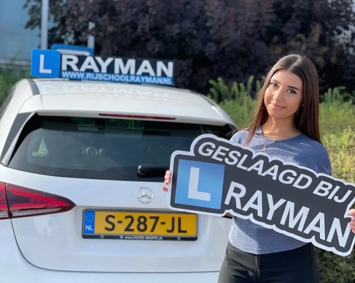 Vrouw houdt een bord met de tekst 'Geslaagd bij Rayman' en een L-onderwijsschild naast een witte auto met een Nederlandse kentekenplaat.