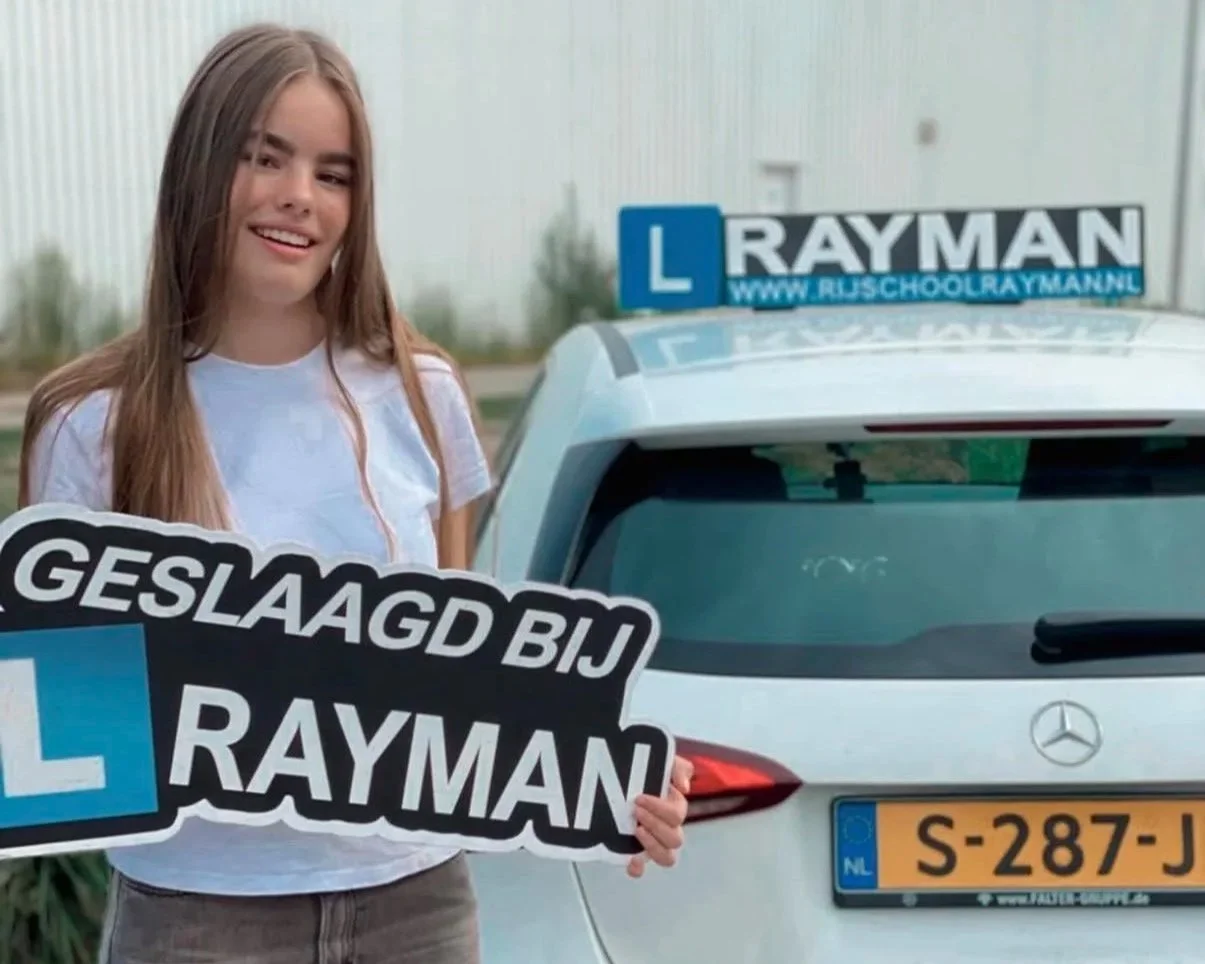 Vrouw met lang bruin haar in witte t-shirt die een bord vasthoudt met de tekst 'Geslaagd bij Rayman', naast een witte auto met een oplegger en een verkeersbord met een 'L' en een websiteadres.