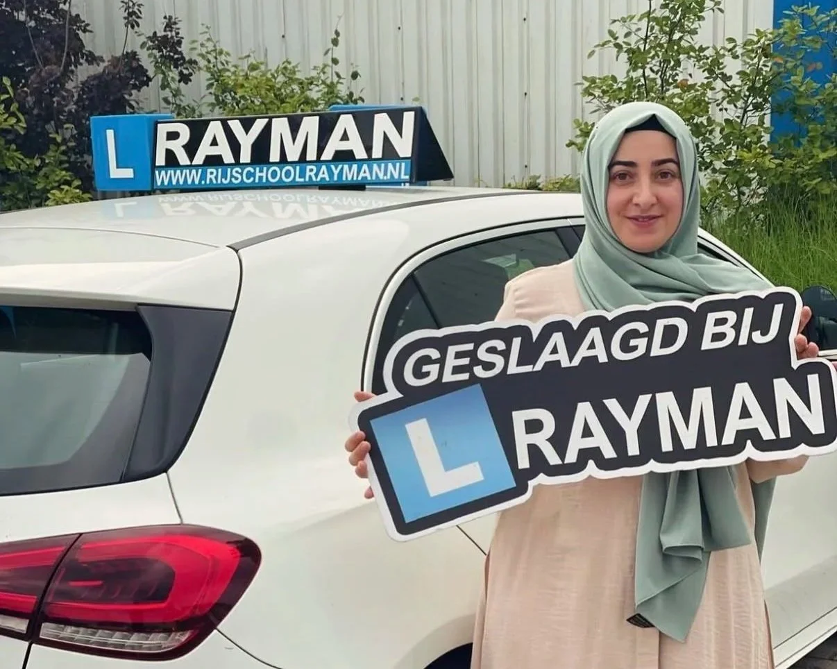 Vrouw met hoofddoek houdt een bord omhoog met de tekst 'GELUKT BIJ RAYMAN' in de buurt van een witte auto met een oplegger die een rijschoolbordje draagt, die online te zien is.