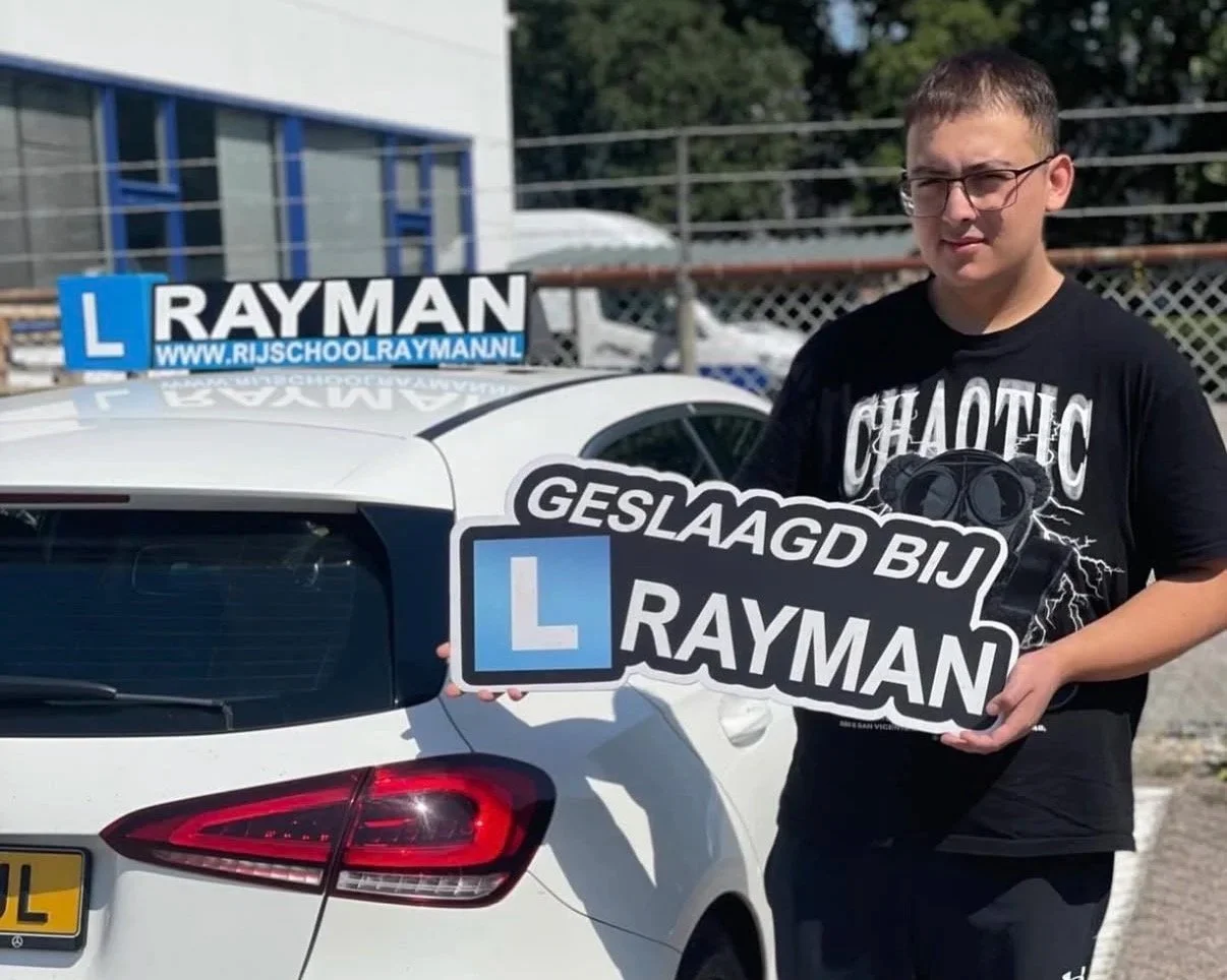 Jonge man met bril staat naast witte auto met lesauto-signaal en houdt een bord met tekst 'Geslaagd bij Rayman' en een L-cursusbord op het dak.