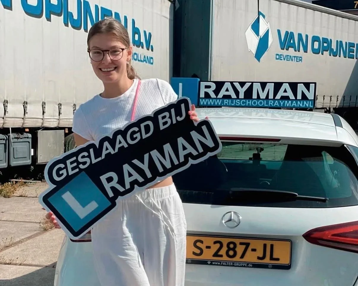 Een vrouw met een grote glimlach houdt een bord vast dat zegt "Geslaagd bij Rayman" en een rijbewijs, standend naast een witte Mercedes met een Nederlands kenteken, voor een vrachtwagen met bedrijfslogo's Van Opijnen en Rayman.