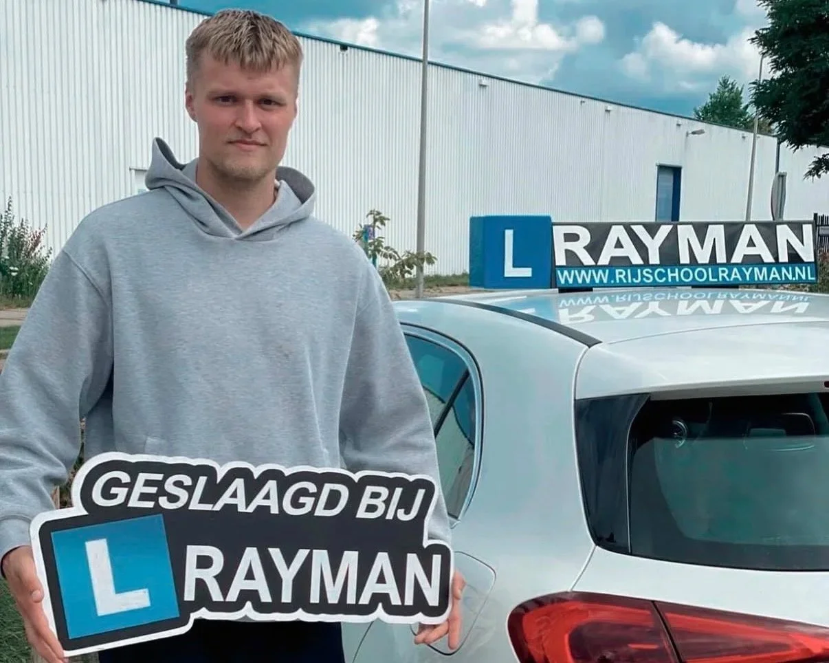 Een jonge man staat naast een auto, die een L-plate en een banner met de tekst 'RAYMAN' en de website 'www.rijschoolrayman.nl' heeft. Hij houdt een bord vast met de tekst 'GELAAAGD BIJ RAYMAN' en een L-plate. De man draagt een grijze hoodie.