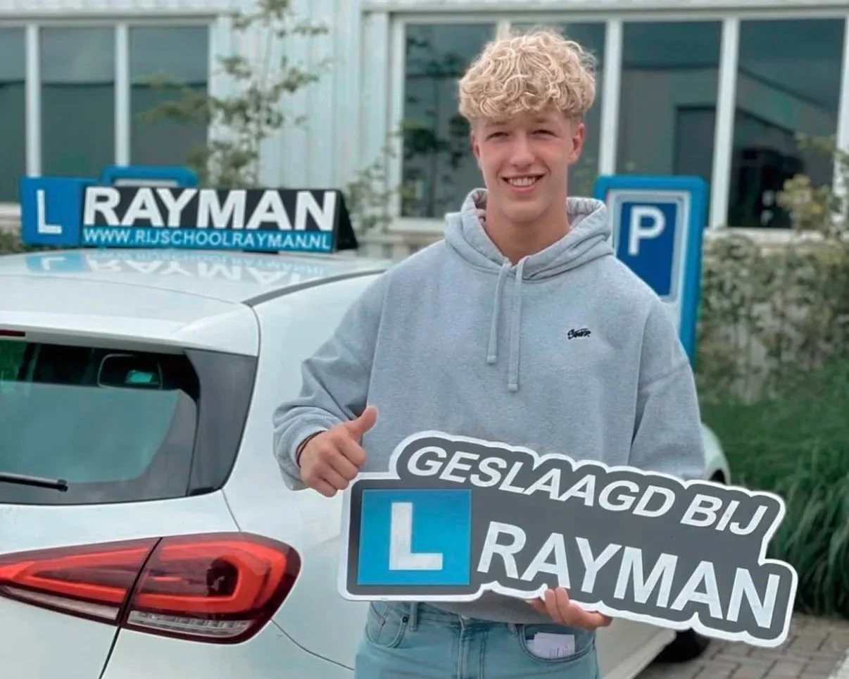 Glücklicher junger Mann steht vor einem Auto mit Fahrschullogo, hält ein Schild mit der Aufschrift «Geslaagd bij Rayman» und zeigt Daumen hoch.
