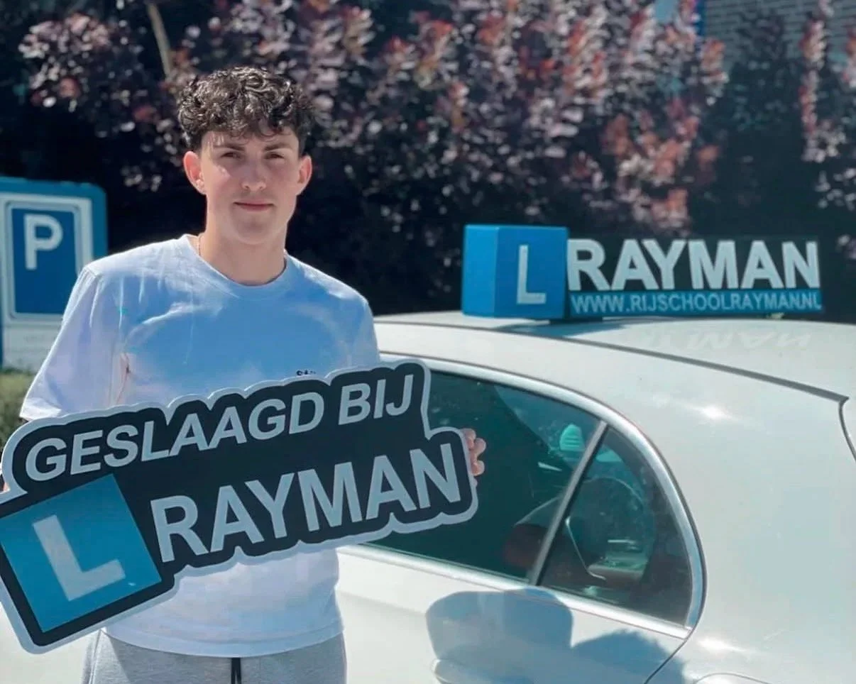 Jonge man poseert met auto en verkeersschoolborden voor rijopleiding, in tuin met bloeiende boom op achtergrond, in Nederland.