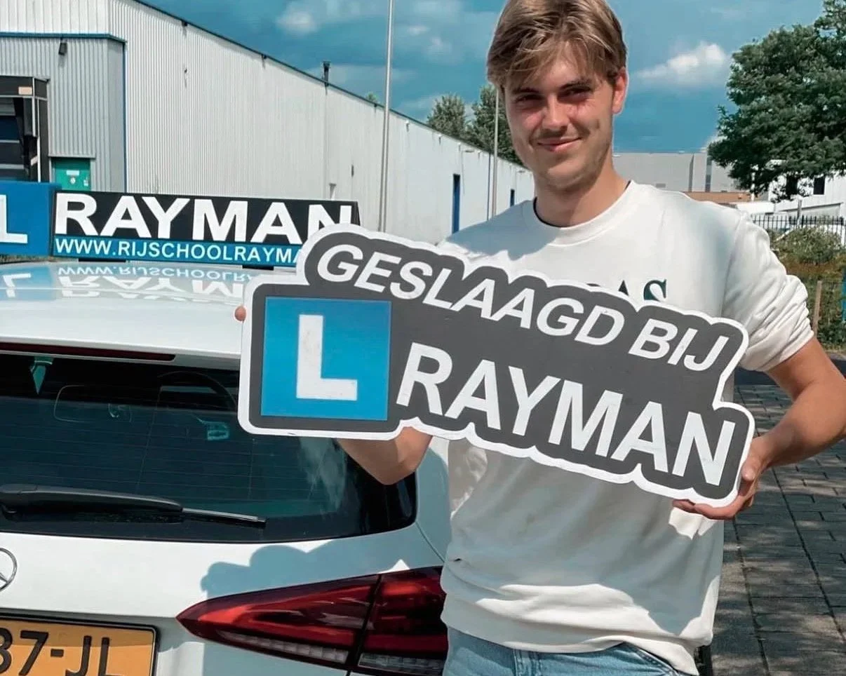 Man houdt een bord met de tekst 'GELUKKIG GESLAAGD BIJ RAYMAN' en een opleidingsauto met verkeersborden in de achtergrond.