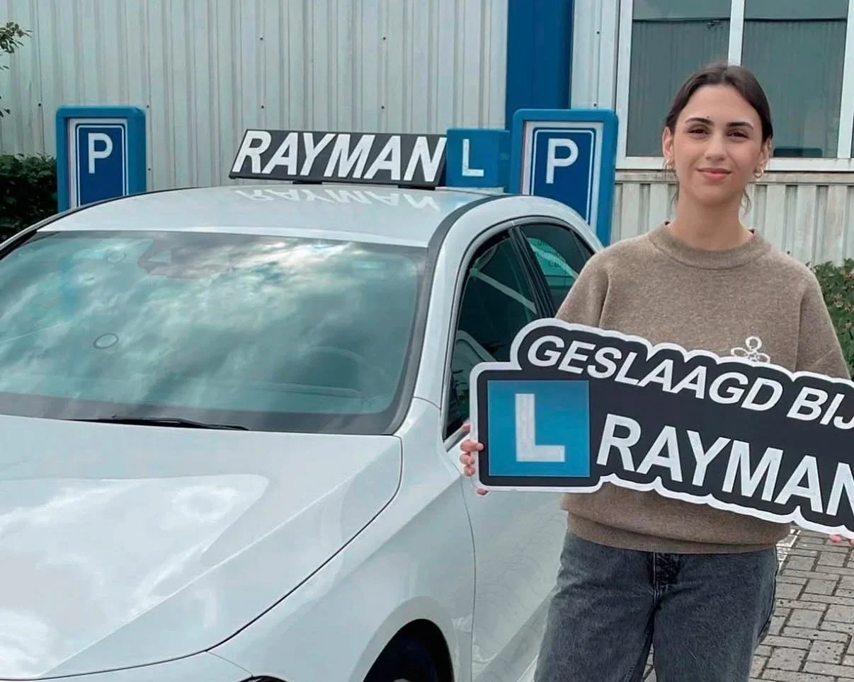 Vrouw staat naast een witte auto met verkeersborden en tekenen voor rijopleiding, houdt een bord met de tekst 'Geslaagd bij Rayman'.