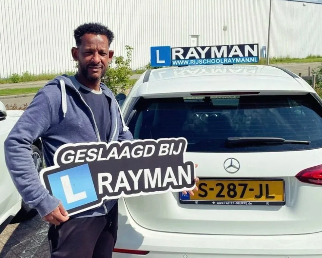 Man houdt bord met tekst 'GESLAAGD BIJ RAYMAN' voor witte Mercedes met Nederlandse kentekenplaat, achter auto bevindt zich een bord met de naam 'RAYMAN' en een L-plate.
