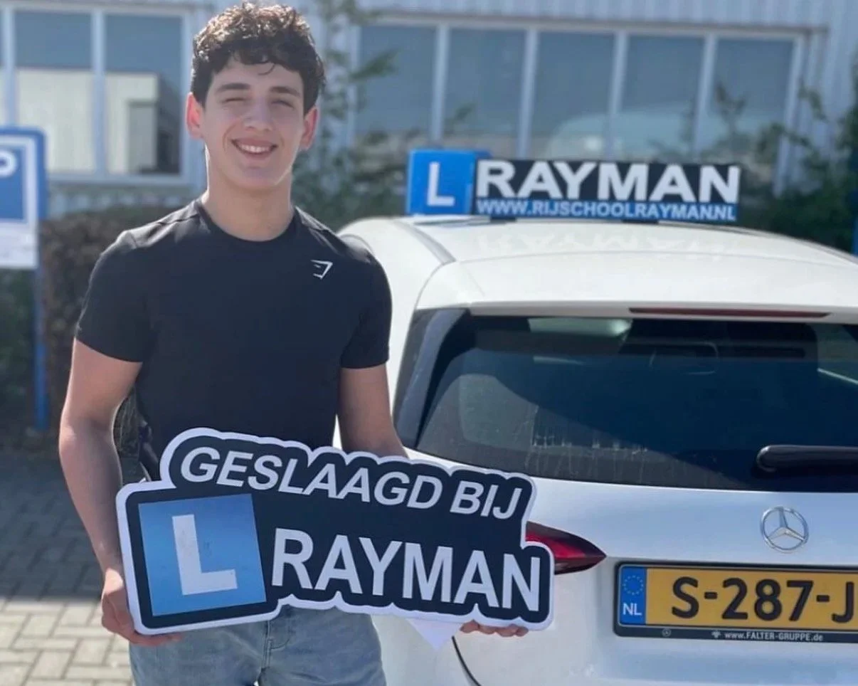 Een jonge man staat voor een witte auto met een L-plate en houdt een bord vast dat zegt 'Geslaagd bij Rayman'.