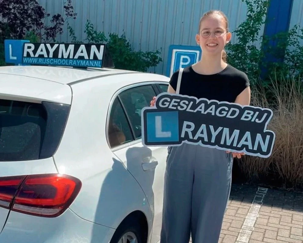 Vrouw met een balk waarop staat 'Geslaagd bij Rayman' voor haar auto met een 'L' lesbord en een 'www.rijschoolrayman.nl' banner.
