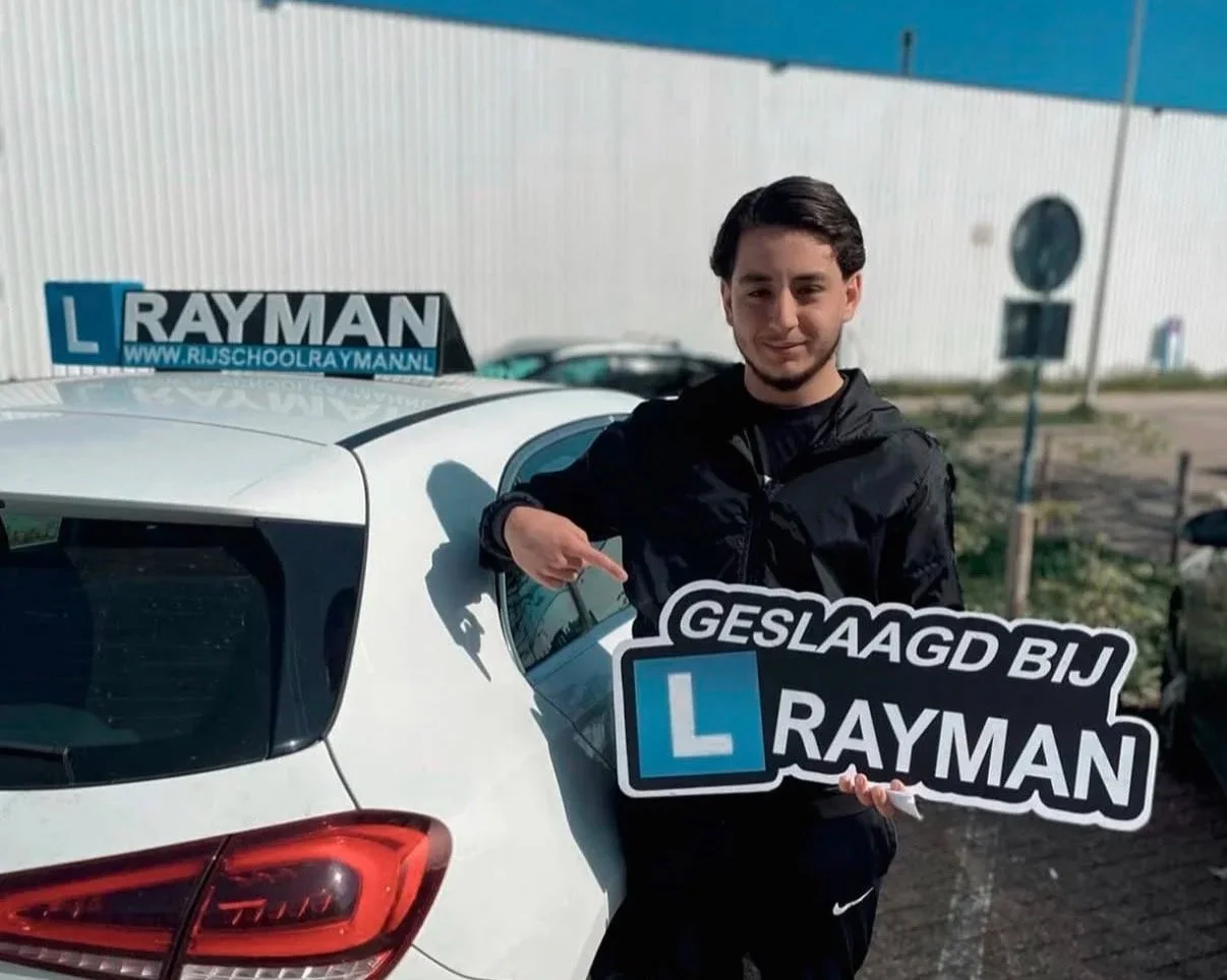 Een man met kort zwart haar die voor een witte auto staat, een verkeersschoolauto, en een bord vasthoudt met de tekst 'GELAAGD BIJ RAYMAN' en een 'L' verkeersbord, terwijl hij naar de camera wijst.
