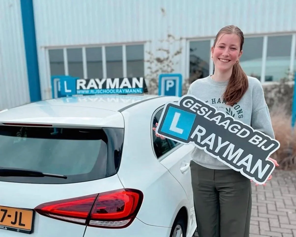 Vrouw die glimlachend naast haar witte auto staat, terwijl ze een bord vasthoudt dat zegt 'GESLAAGD BIJ RAYMAN' en een ander bord met 'L RAYMAN' op de auto ligt.