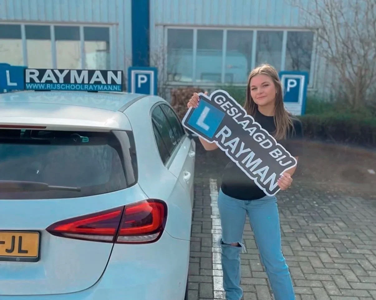 Een dame uit Twello die is geslaagd met bord geslaagd bij Rijschool Rayman in haar handen, voor de Mercedes lesauto van Rijschool Rayman