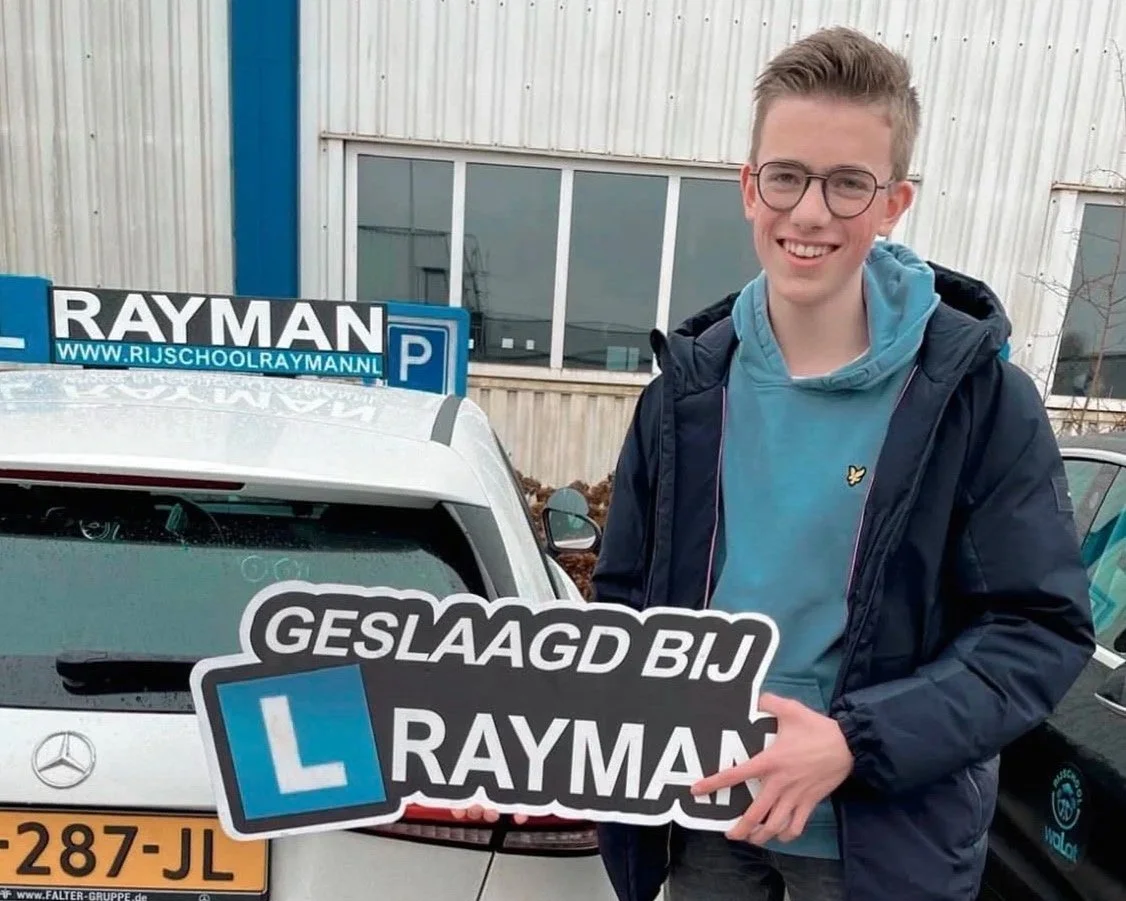 Jongen die poseert naast een auto met verkeerslesbord, holding een bord met de tekst 'Geslaagd bij Rayman' en een grote L voor leerrijbewijs.