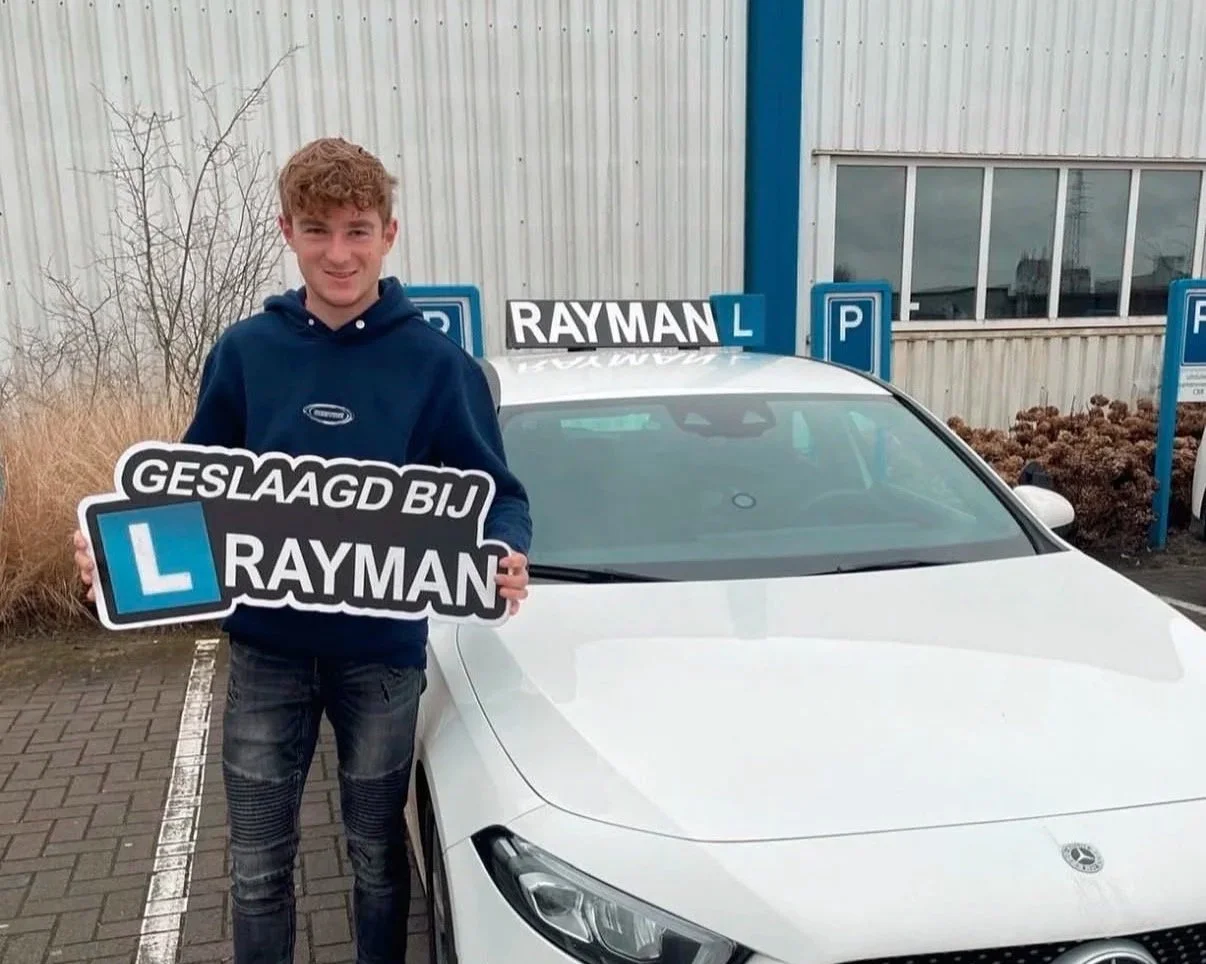 Een jonge man staat naast een witte auto met een bord in zijn handen dat zegt 'Geslaagd bij Rayman' en een L-helmteken, met het Rayman rijexamencentrum op de achtergrond.