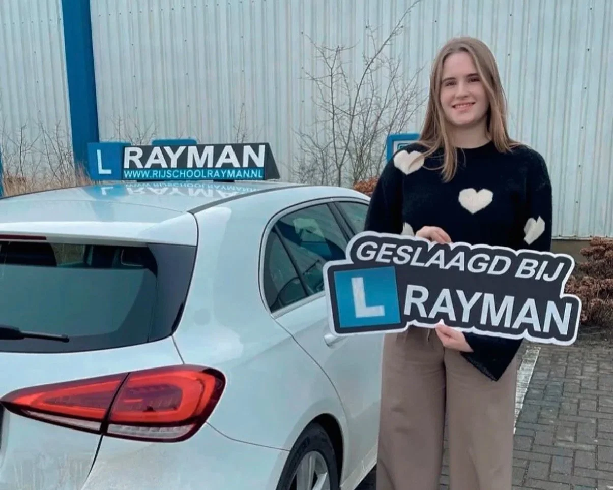 Een jonge vrouw staat naast een witte auto met een leerlingchauffeursbord op het dak. Ze houdt een bord vast dat zegt 'Geslaagd bij Rayman'.