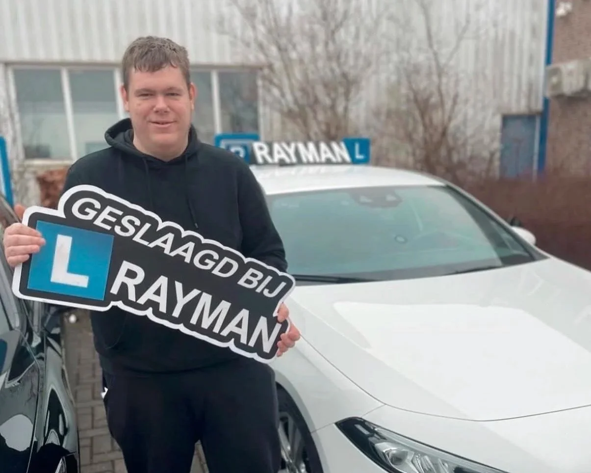 Man houdt een bord vast met de tekst 'GELAGD BIJ RAYMAN' en een blauwe 'L' erop, terwijl hij voor een witte auto staat met een rijschoolbord op het dak.