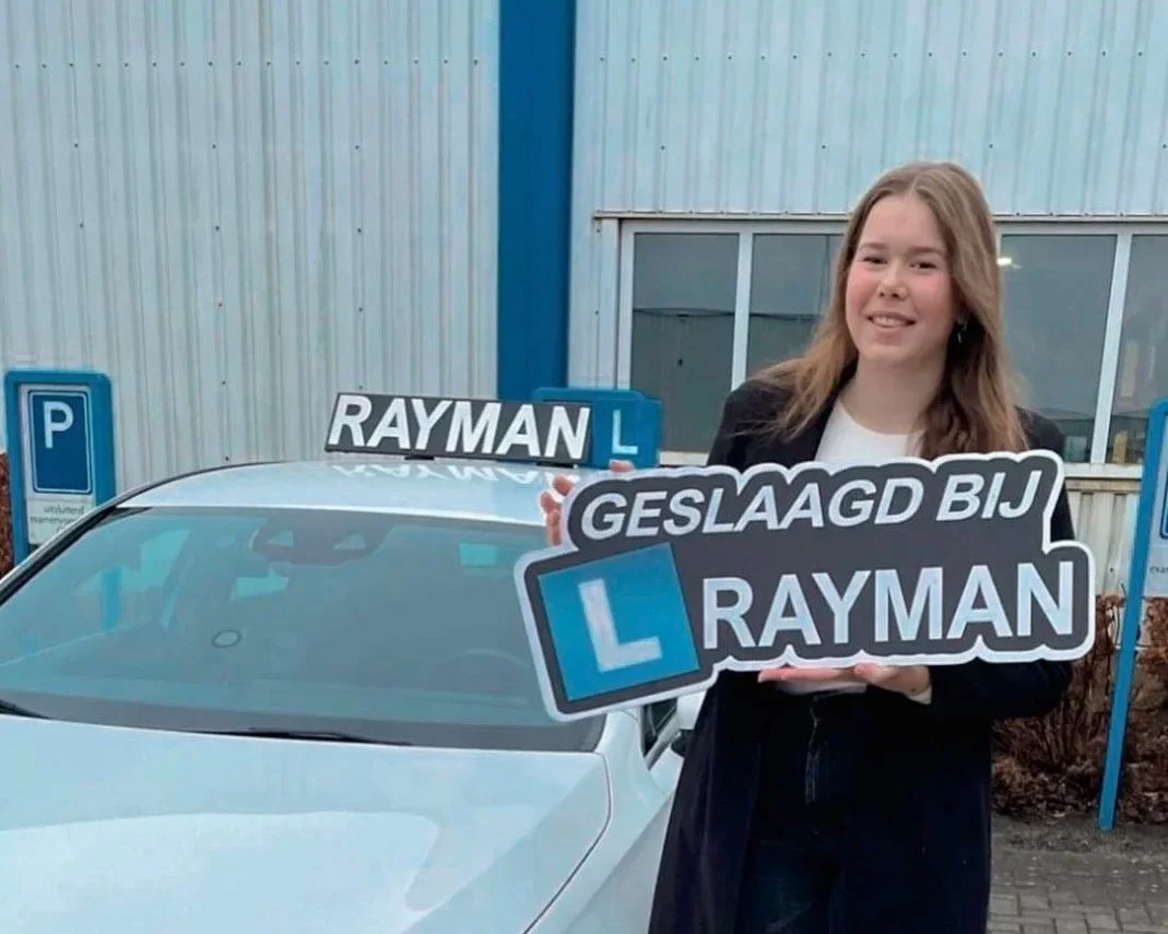 Een vrouw houdt een bord met de tekst 'GESlaagd BIJ RAYMAN' voor haar, terwijl ze naast een auto met een L-plate zit. Er zijn blauwe parkeergelegenheden op de achtergrond.