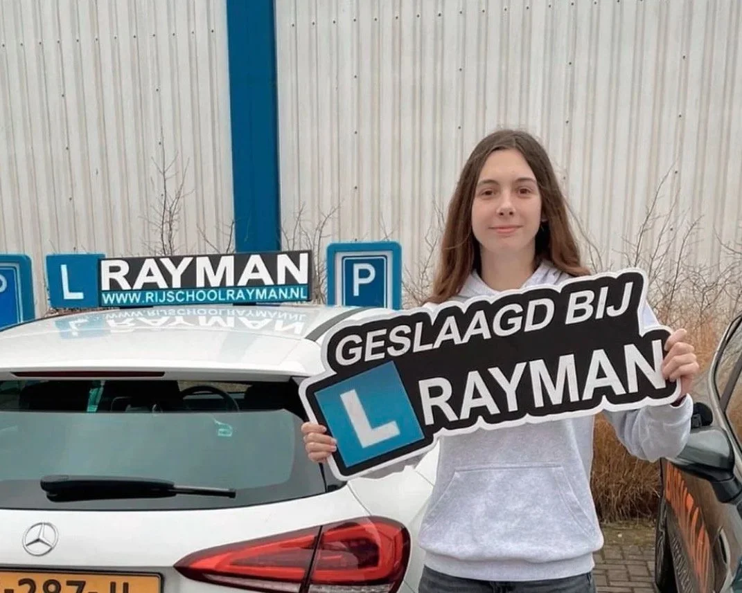 Meisje houdt een bord vast dat zegt 'Geslaagd bij Rayman' voor een rijden-schoolauto met borden op het dak, buiten staan er bomen en een metalen muur.