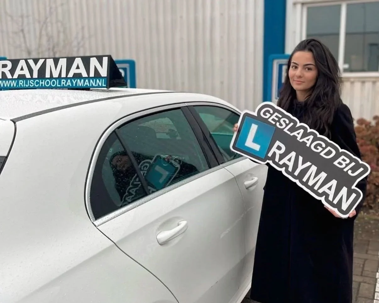 Vrouw met lang donker haar en zwarte jas, die een bord vasthoudt met tekst 'GESLAAGD BUI RAYMAN', naast een witte auto met een rijschoolbord op het dak. De rijschoolwebsite 'www.rijschoolrayman.nl' is zichtbaar.
