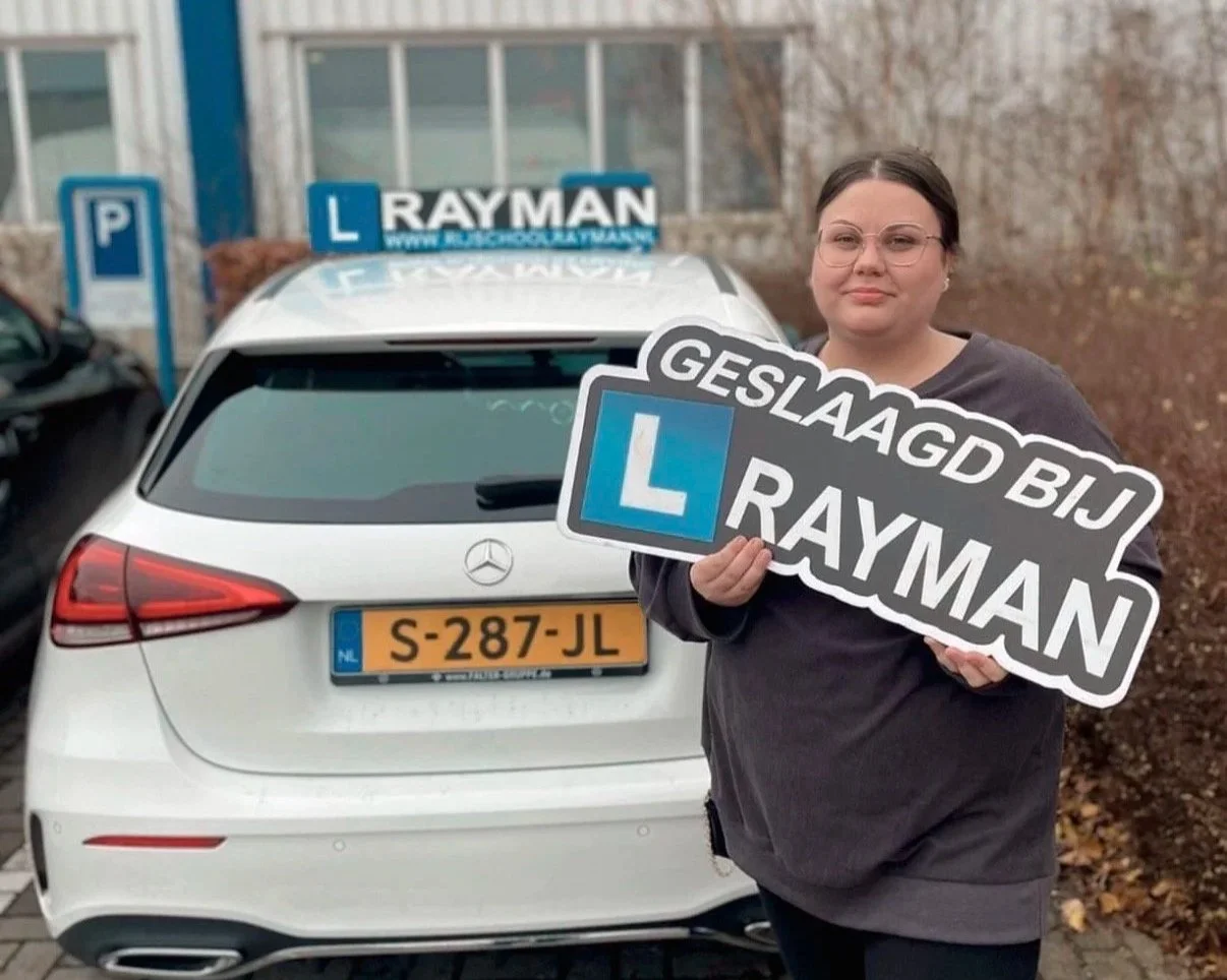 Vrouw met een bord dat zegt 'Geslaagd bij Rayman' voor een witte auto met een oplegger, het verkeerbord met haar opleiding hangt op het dak van de auto.