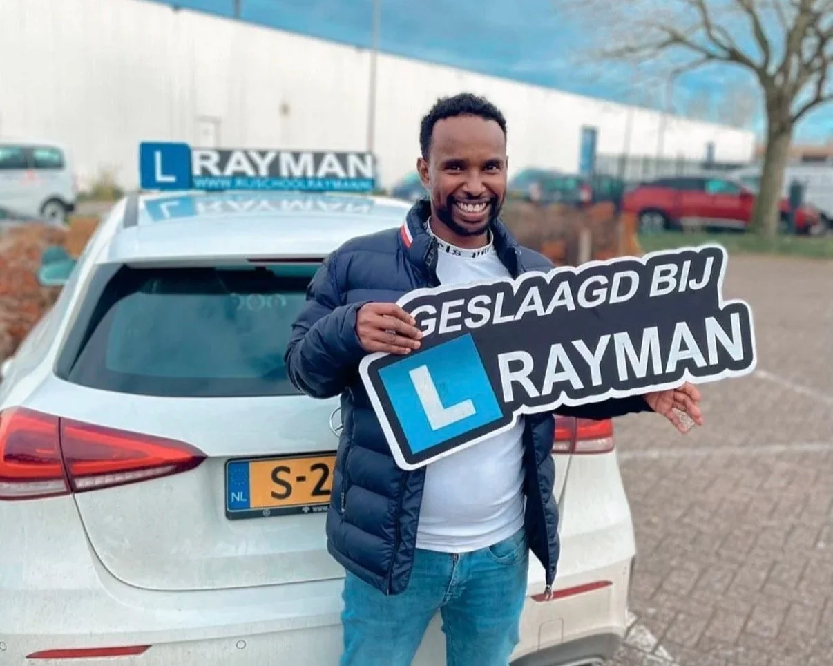 Man staat voor een witte lesauto met een L-ighaalbord, hij houdt een bord met tekst "Geslaagd bij Rayman" en lacht, terwijl hij zich voorbereidt op rijlessen.