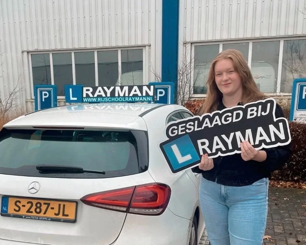 Een jonge vrouw in een zwarte jas en lichte spijkerbroek staat naast een witte auto met rijopleidingssignalering en een bord die zegt 'GESLAAGD BIJ RAYMAN'.