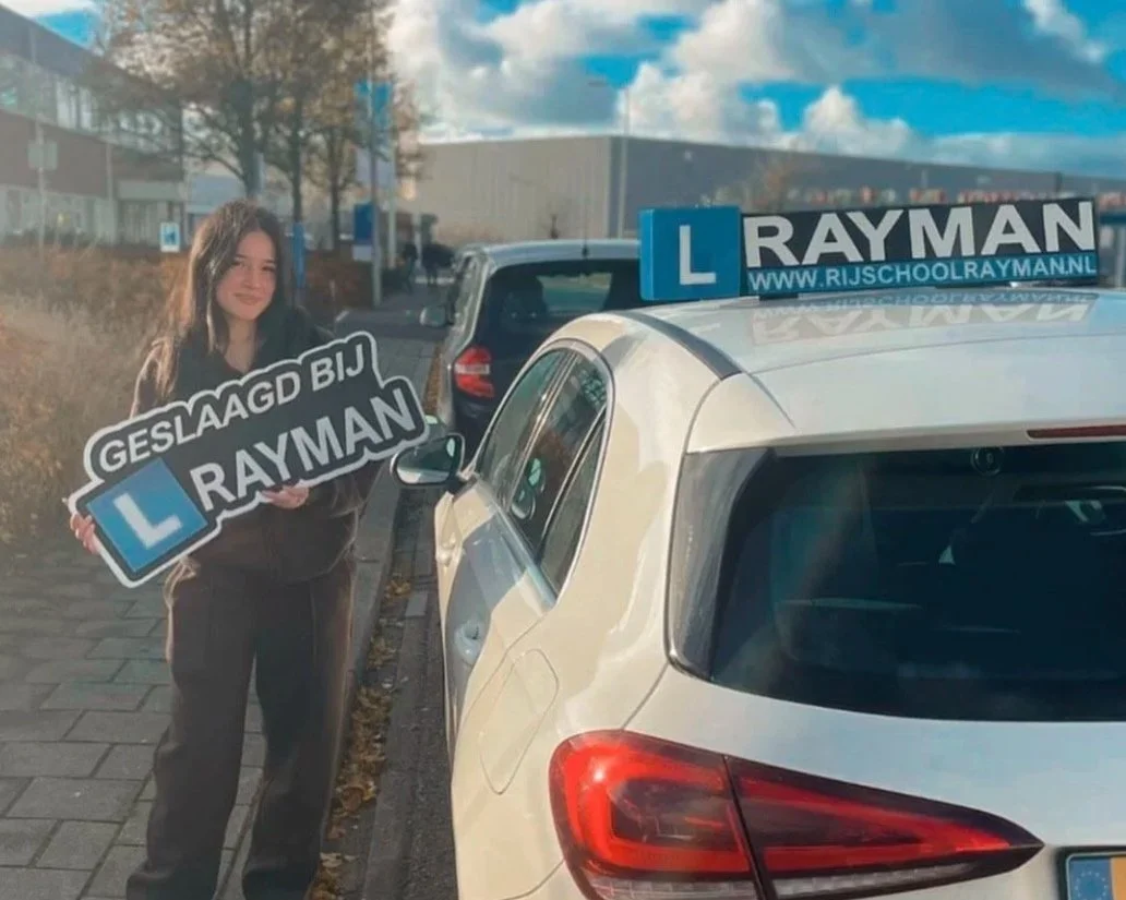 Een jonge vrouw staat naast een witte auto met een instructiebord, terwijl ze een bord vasthoudt dat zegt 'Geslaagd bij Rayman' en een Blue L-lesbord op de auto met de tekst 'www.rijschoolrayman.nl'.