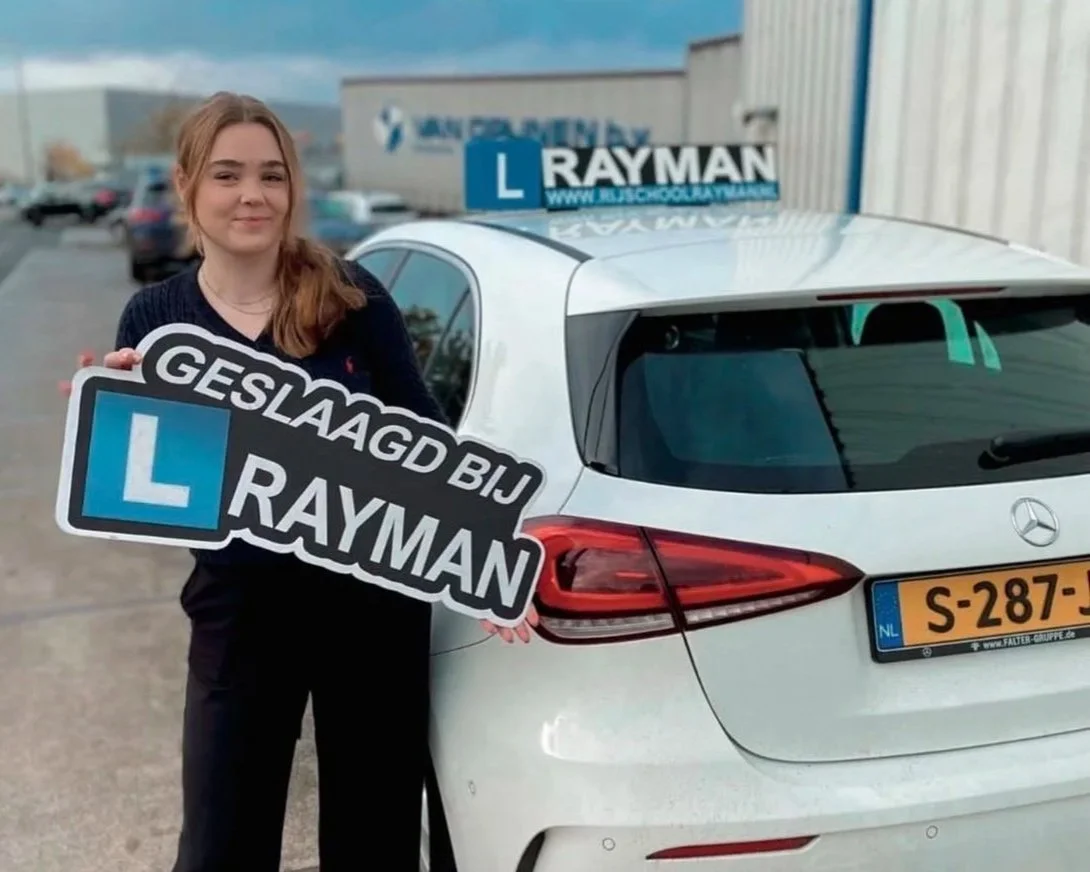 Vrouw met lang haar houdt een bord vasthouden dat zegt 'Geslaagd bij Rayman' met een L-oplader symbool, staand naast een witte auto met een Rayman-slogan op de spiegel en een Nederlands kenteken.