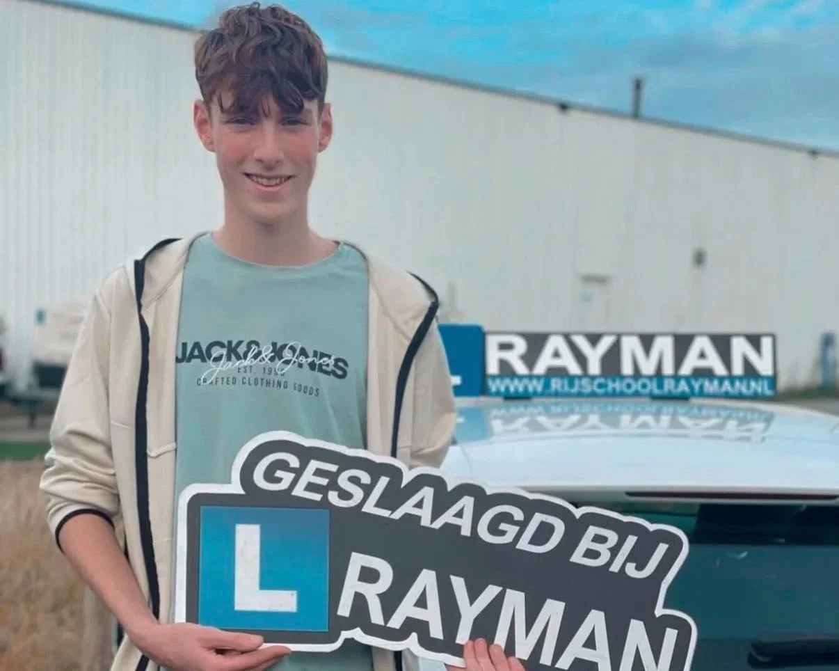 Een jonge man staat buiten voor een wit gebouw met een blauwe hemel op de achtergrond, hij houdt een bord vast dat zegt 'Geslaagd bij Rayman' met een grote blauwe 'L'-bord en een gelakte auto ernaast.