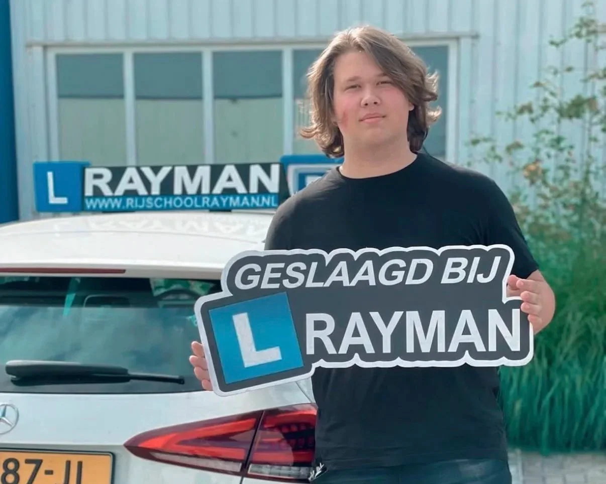 Een jongen met halflang haar die een bord vasthoudt met de tekst 'Geslaagd bij Rayman' en een L-plate, buiten voor een auto met een Rayman-signdisplay op de achtergrond.