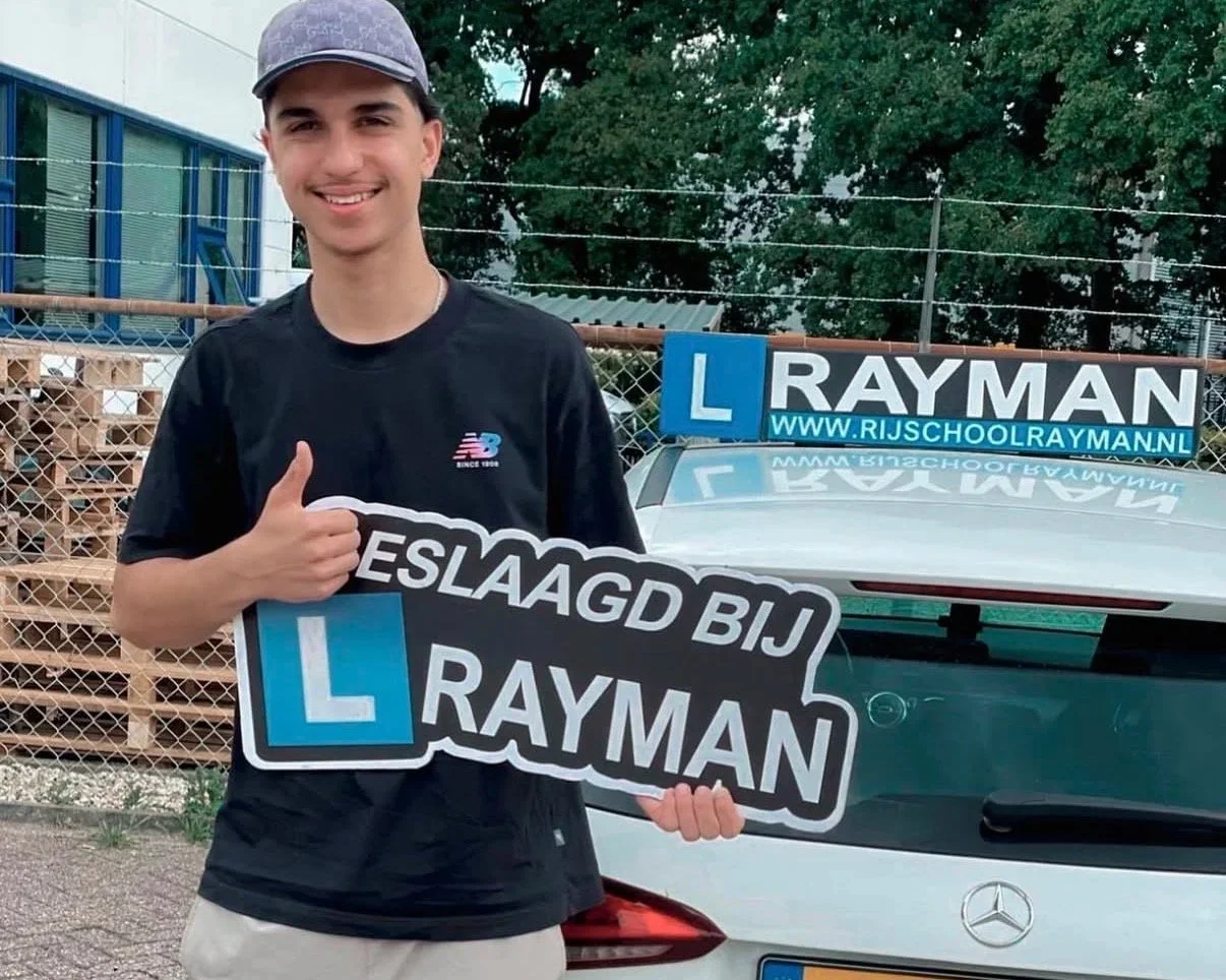 Jonge man glimlachend naast een auto met een L-plate en een bord dat zegt 'Geslaagd bij Rayman'.
