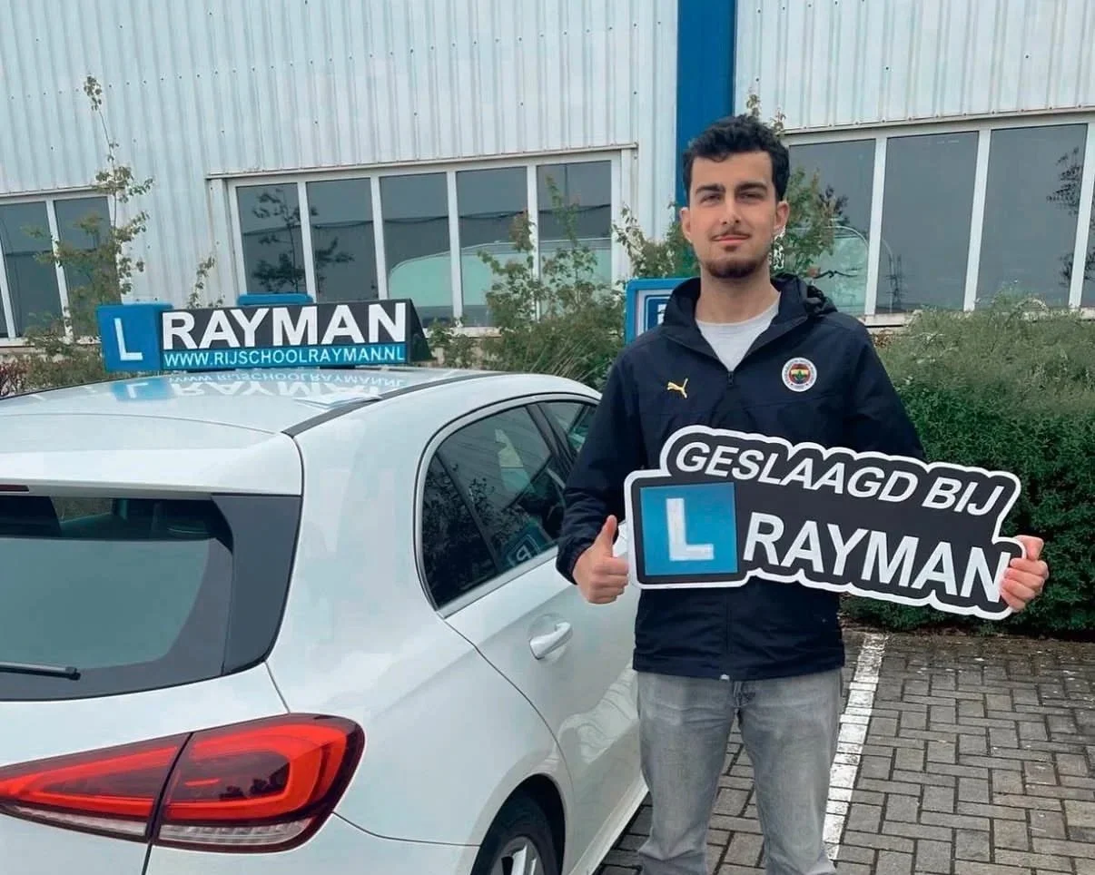 Een jonge man staat naast een witte auto met een L-opleiding en een bord dat zegt 'Geslaagd bij Rayman'. Hij houdt een groot bord met dezelfde tekst en een L-opleiding ernaast, terwijl hij een duim omhoog geeft.