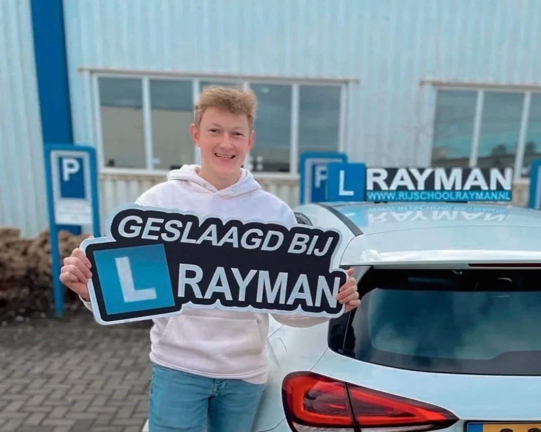 Jonge man met kort haar die een bord vasthoudt met de tekst 'GESLAAGD BIJ RAYMAN' en een L- candidate achter een auto, bij een rijschool. Dus, waarschijnlijk is hij net geslaagd voor zijn rijexamen.