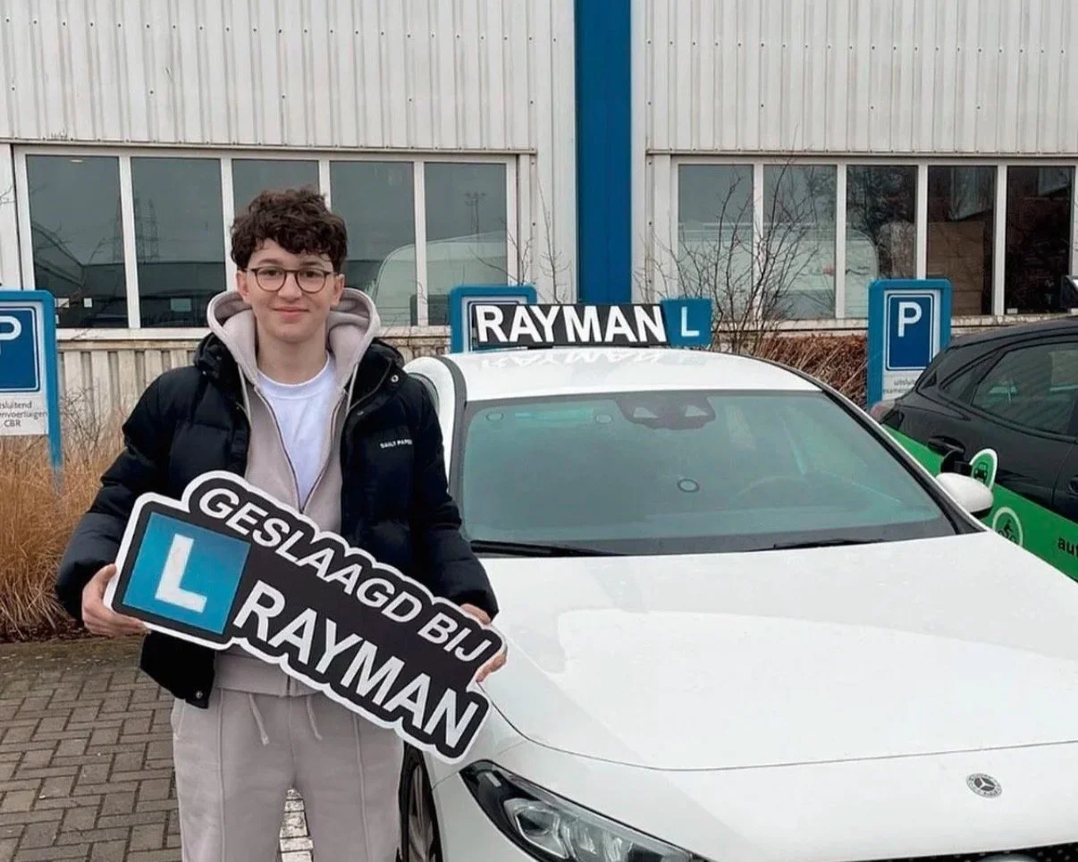 Een jongeman staat voor een witte auto met een 'RAYMAN' rijlesbord op het dak, hij houdt een bord met de tekst 'GESLAAGD BU RAYMAN' en een L. Hij staat op een parkeerplaats met parkeermeters en gebouwen achter hem.