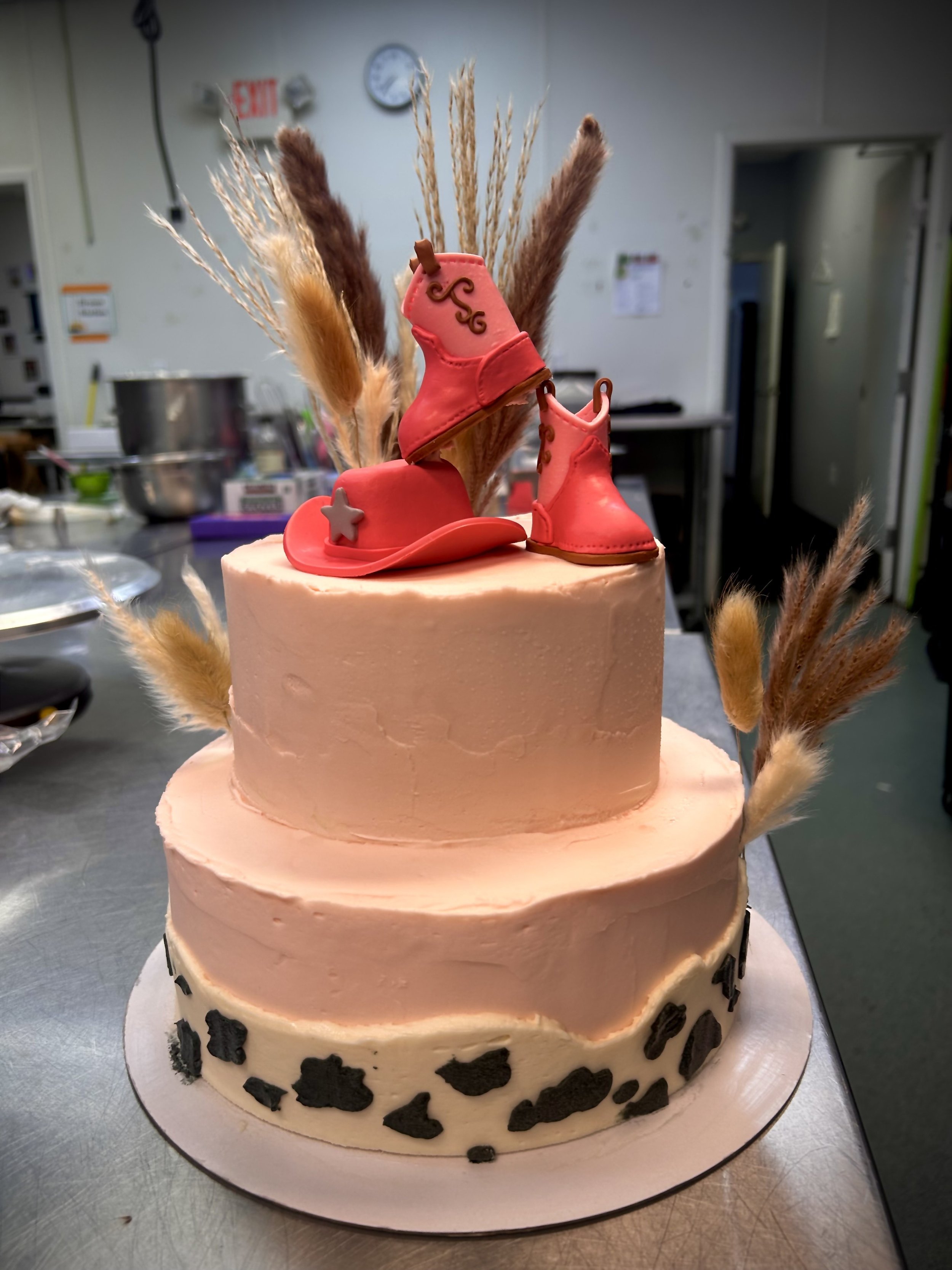 A two-tiered cake decorated with pink icing, featuring cowboy-themed cake toppers including a cowboy hat, boots, and a star, with dried grass and pampas grass accents.
