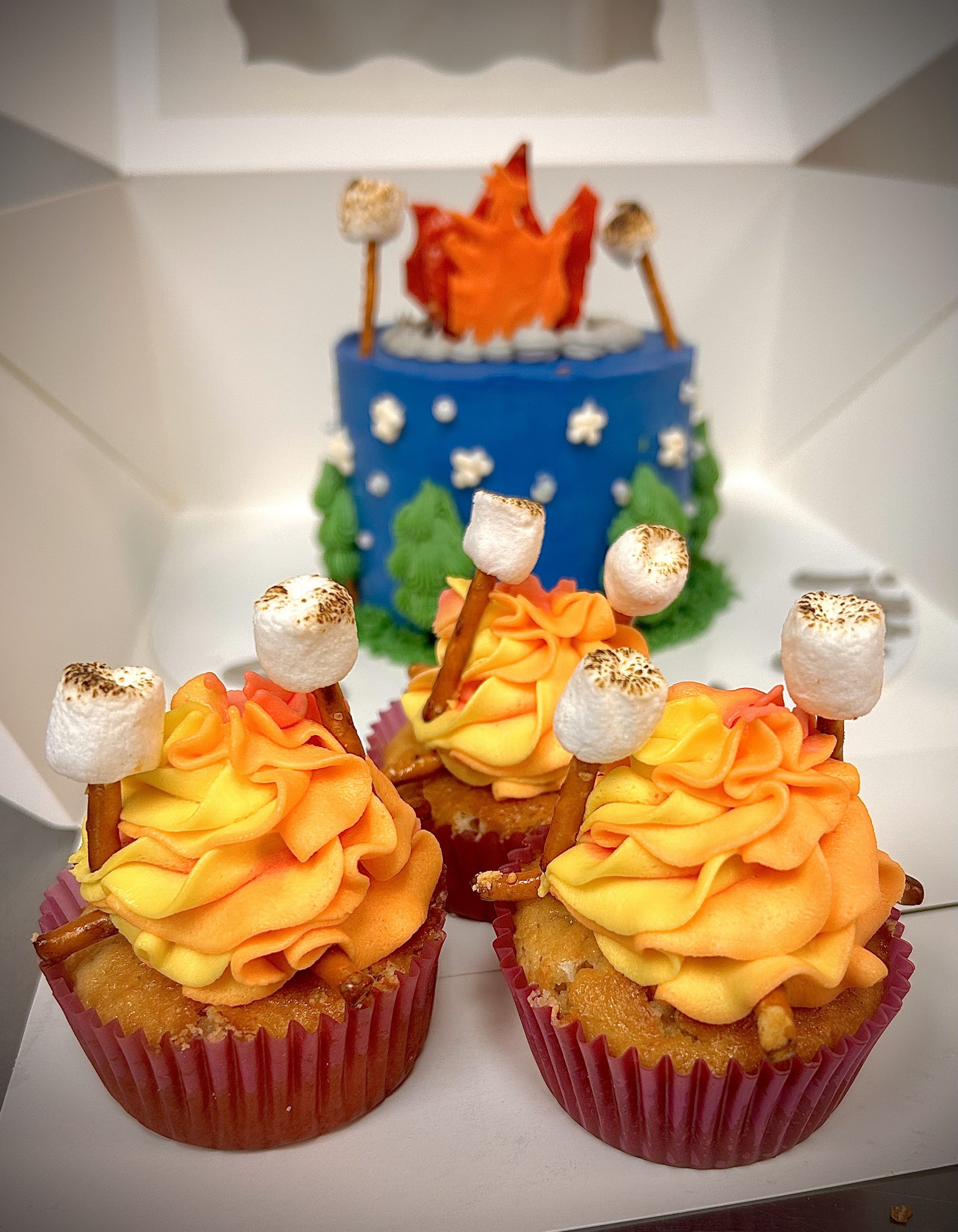 Three cupcakes decorated with yellow and orange frosting and mini pretzel sticks topped with marshmallows in front of a blue cake with green trees and white stars with a red and orange flame decoration on top.