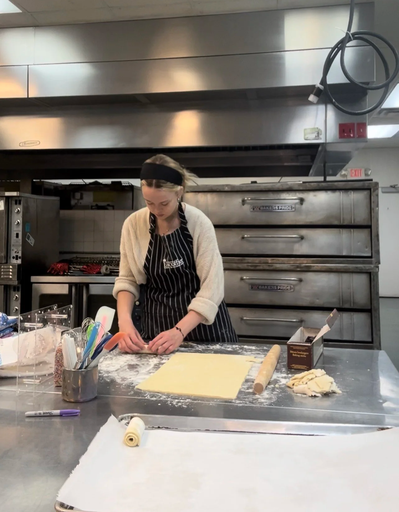 A woman in a black apron is preparing pastry dough on a floured stainless steel countertop in a commercial kitchen. She is rolling out dough with a rolling pin, surrounded by baking tools and ingredients.