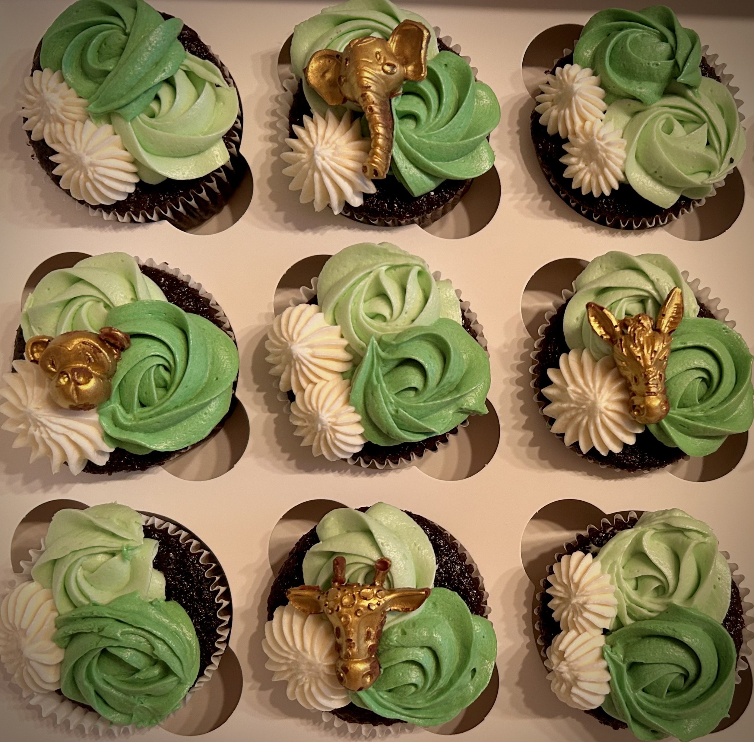 Nine decorated cupcakes with green and white frosting, each topped with a gold animal or animal-themed decoration, including an elephant, giraffe, lion, and zebra, in a 3x3 grid in a white cupcake box.