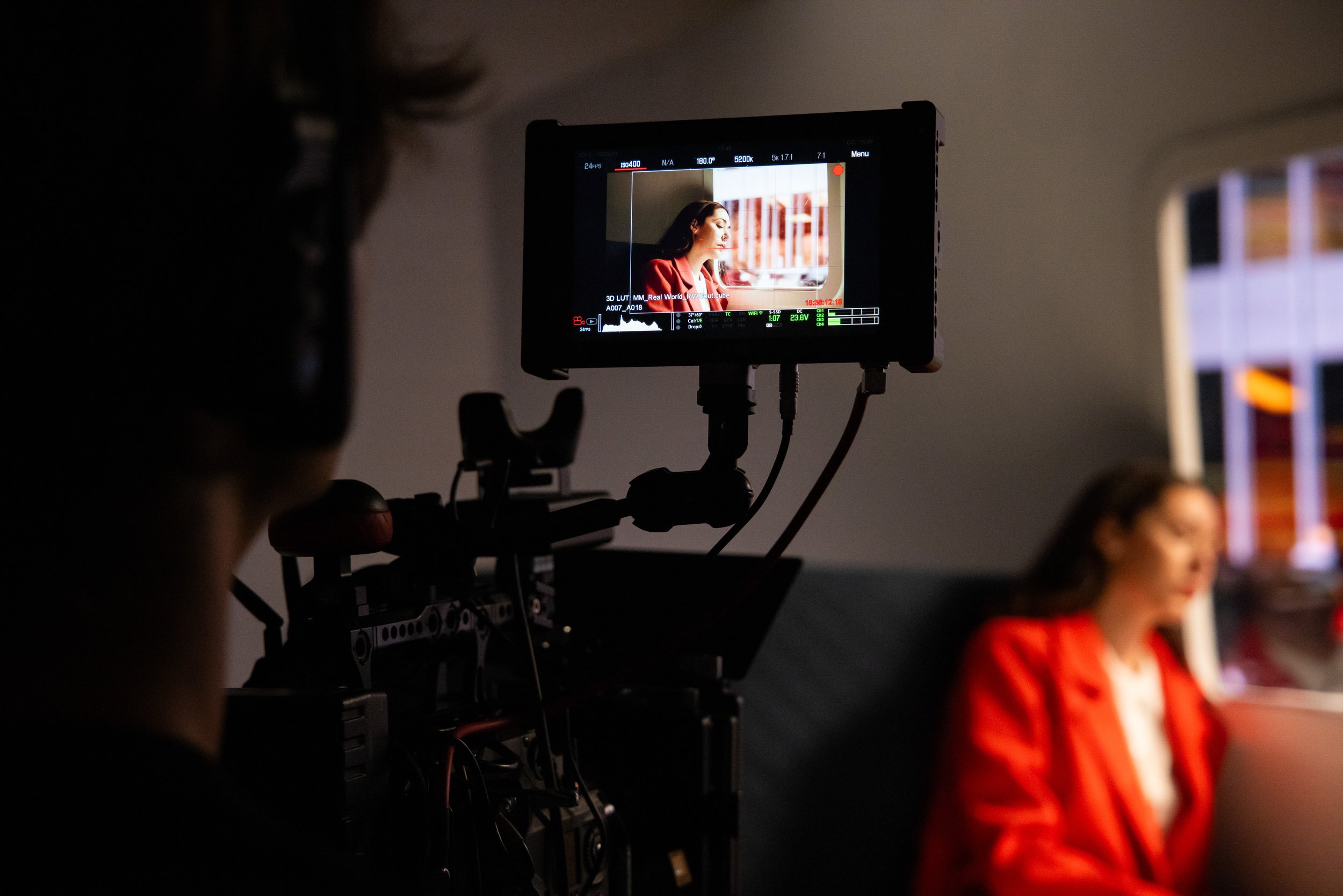 A woman in a red jacket sits in front of a window, being filmed by a camera on a rig, with the camera's external monitor showing her face and profile.