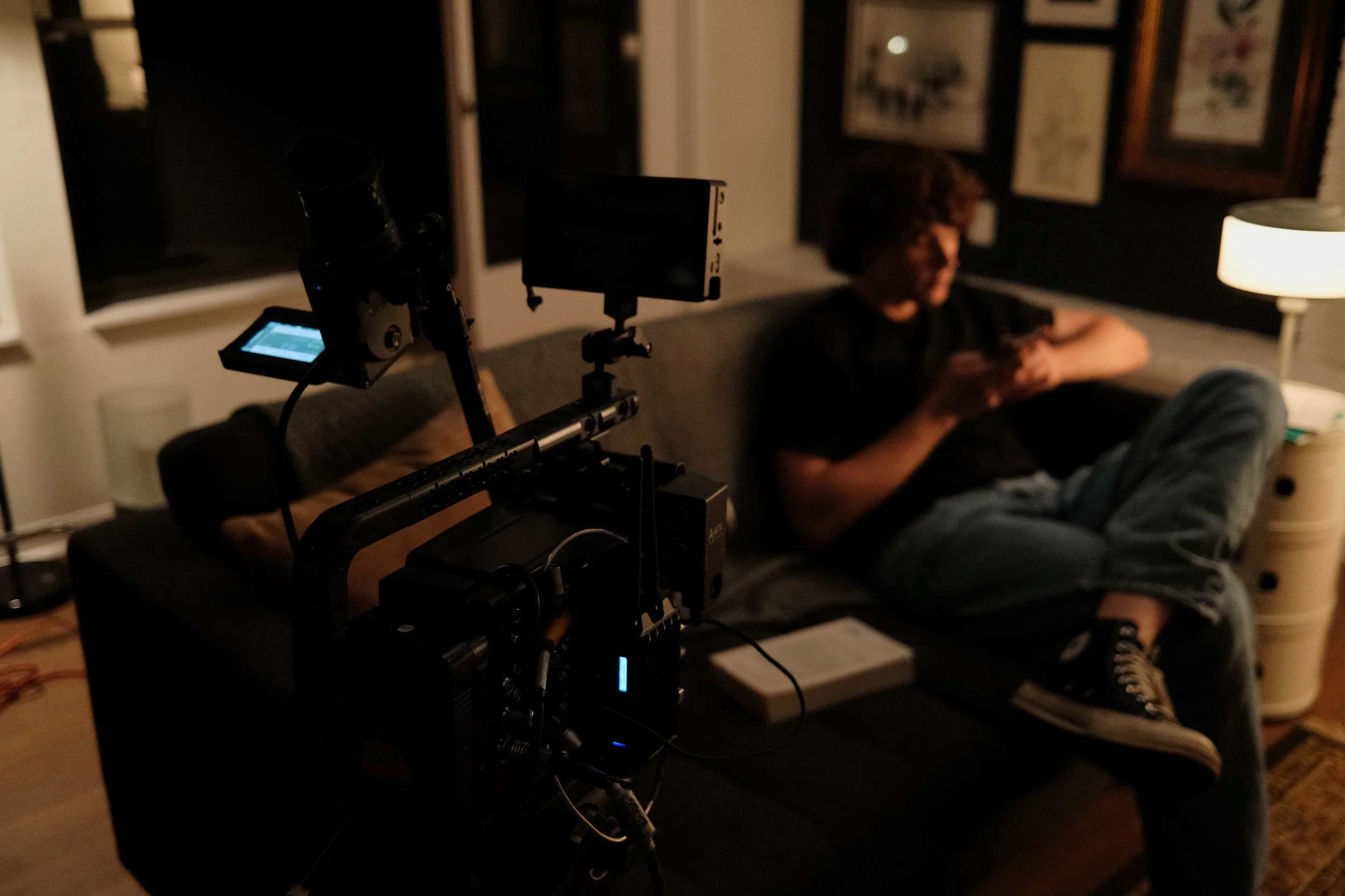 A person is sitting on a couch in a dimly lit room using a phone. A film camera setup is in the foreground, pointing towards the person.