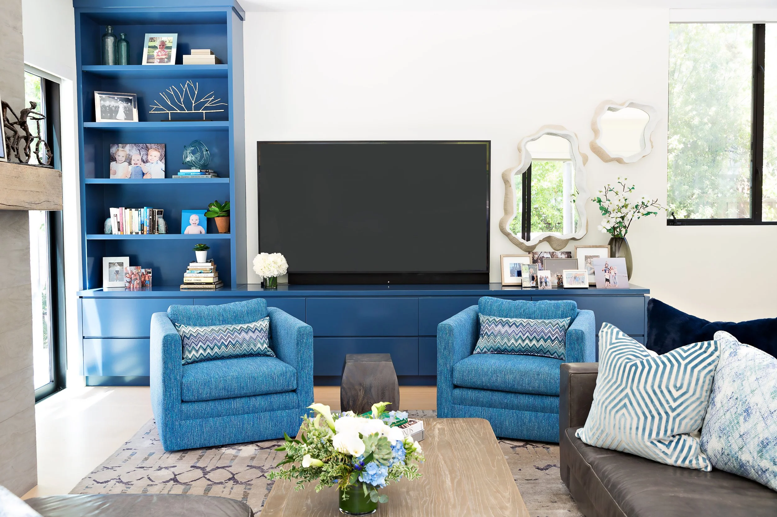 Living room with blue armchairs, a blue bookshelf, a large flat-screen TV, framed photos, decorative mirrors, a window with greenery outside, and a coffee table with a flower arrangement.