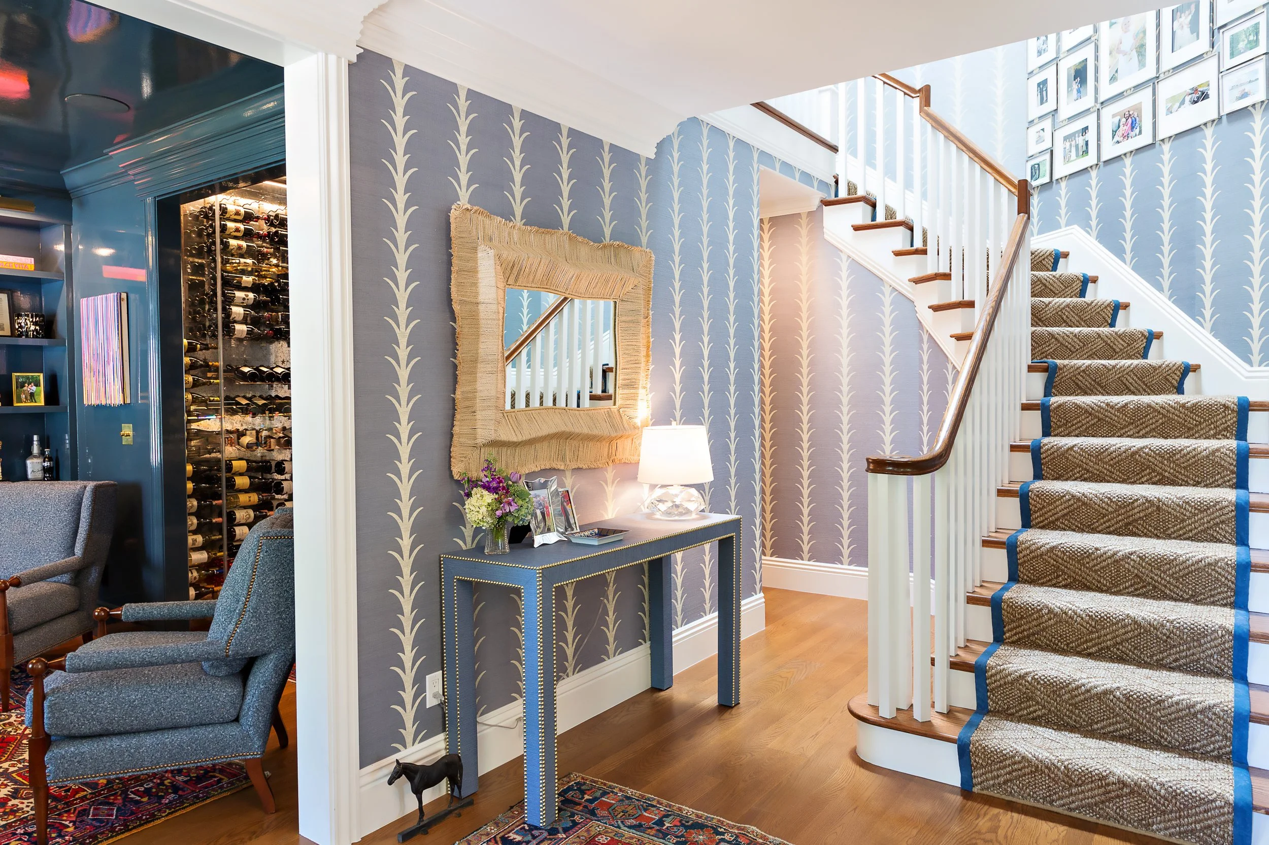 Entryway with a staircase, blue patterned wallpaper, a mirror with a woven frame, a console table with a lamp and flowers, and a wine storage. Part of a living area with armchairs and a bookcase.